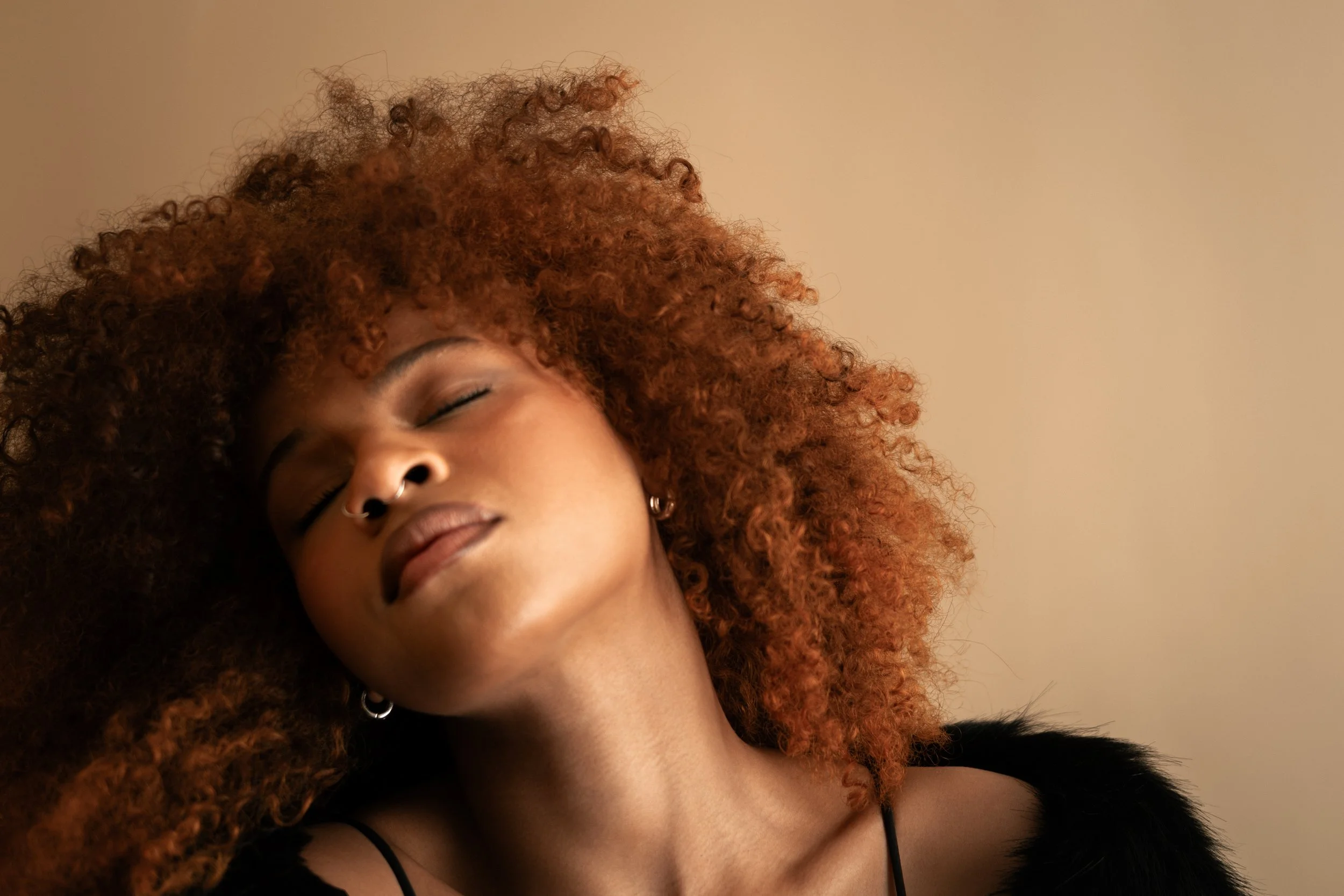 A woman with curly red hair, closed eyes, and a nose piercing, leaning her head back with a serene expression, wearing a black top with thin straps.