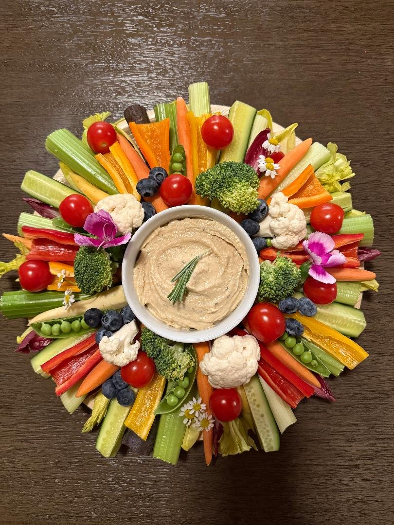 Colorful vegetable platter with cherry tomatoes, celery, carrots, broccoli, cauliflower, blueberries, and edible flowers, centered around a bowl of hummus garnished with rosemary on a brown wooden surface.