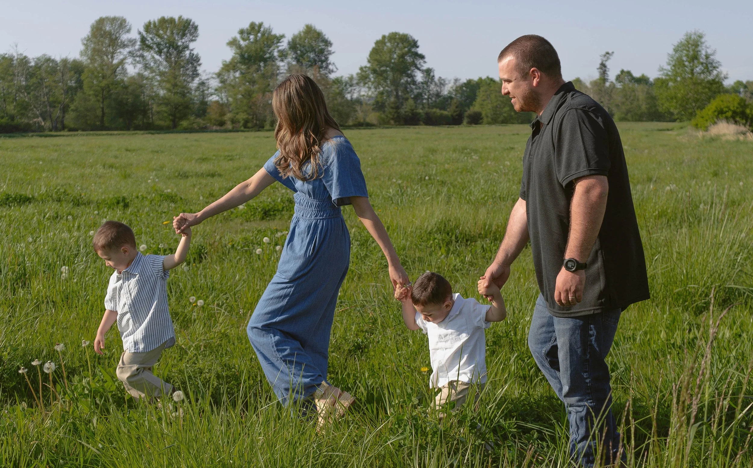A family of four walking in a grassy field on a sunny day, holding hands, surrounded by trees.