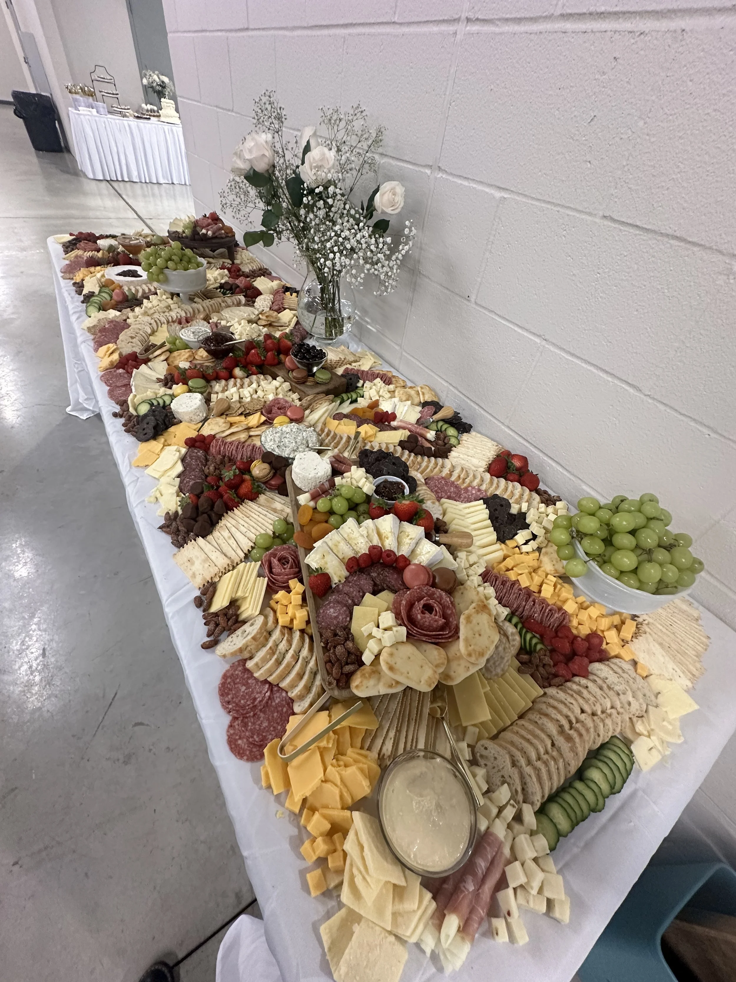 Large cheese and charcuterie spread with assorted cheeses, cured meats, fruits, crackers, and dipping sauces on a long table, decorated with a floral centerpiece of white roses and baby's breath.
