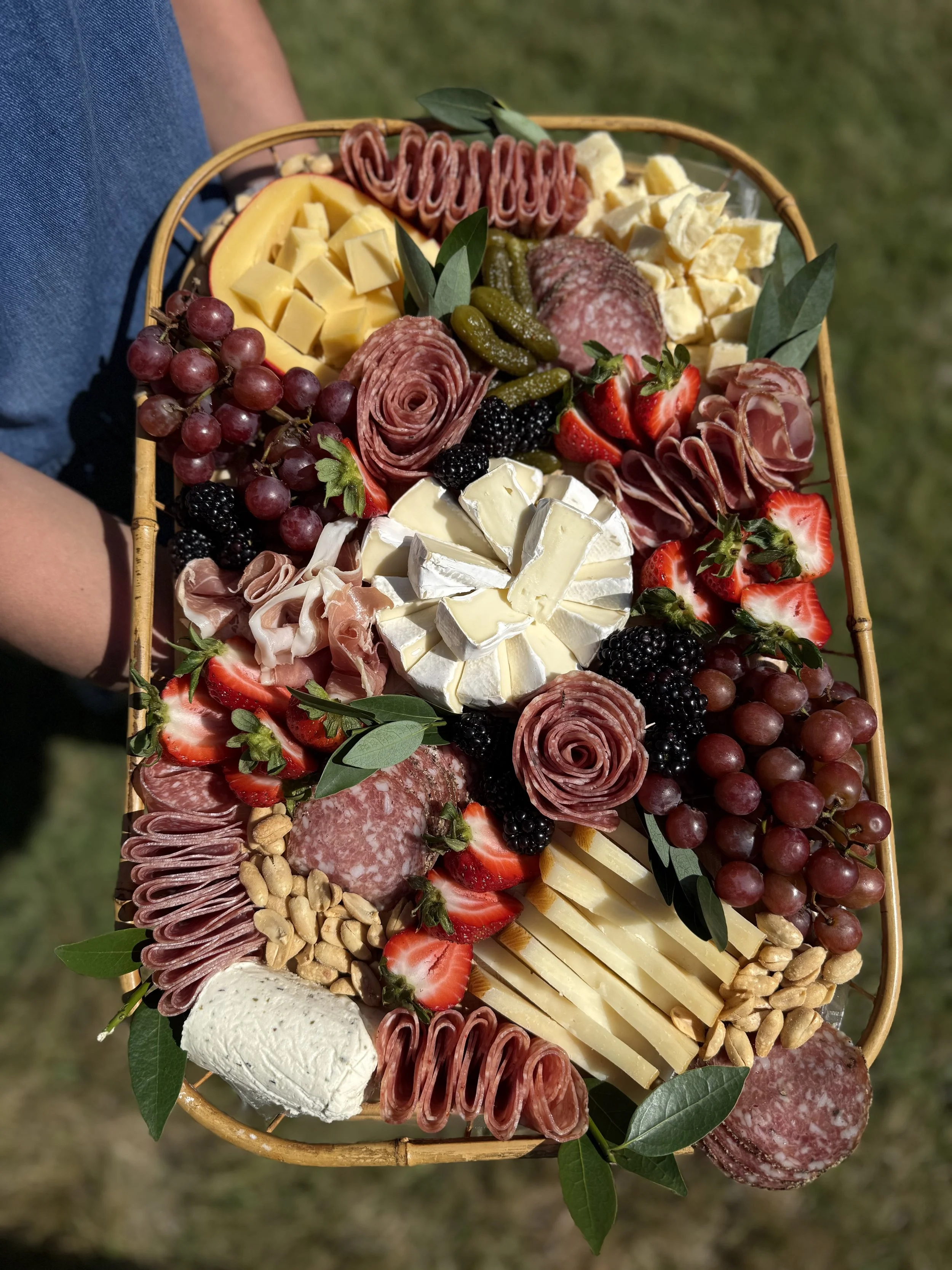 A basket of assorted cheeses, grapes, strawberries, blackberries, dried nuts, cured meats, and green leaves, arranged for a charcuterie platter.