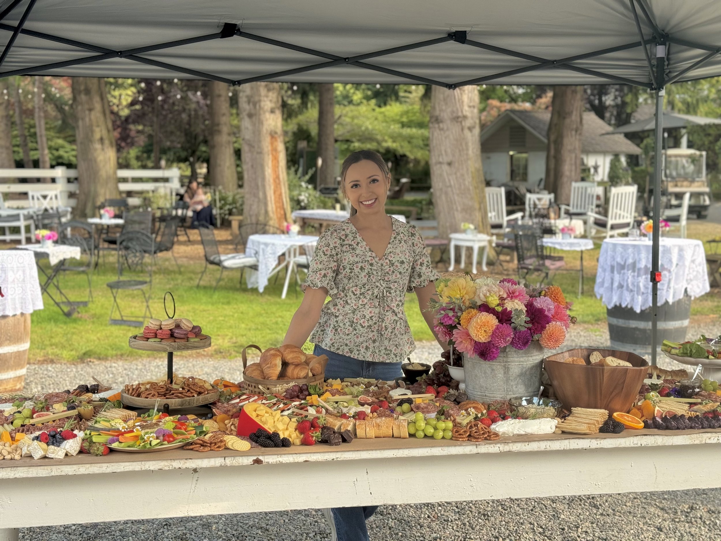 A woman standing behind a table filled with various desserts and snacks at an outdoor event under a canopy. The table has plates of macarons, croissants, cheese, fruits, and other treats. There is a large bouquet of pink, yellow, and orange flowers in a metal bucket on the table. Background shows a garden with trees, white tables and chairs, and people sitting.