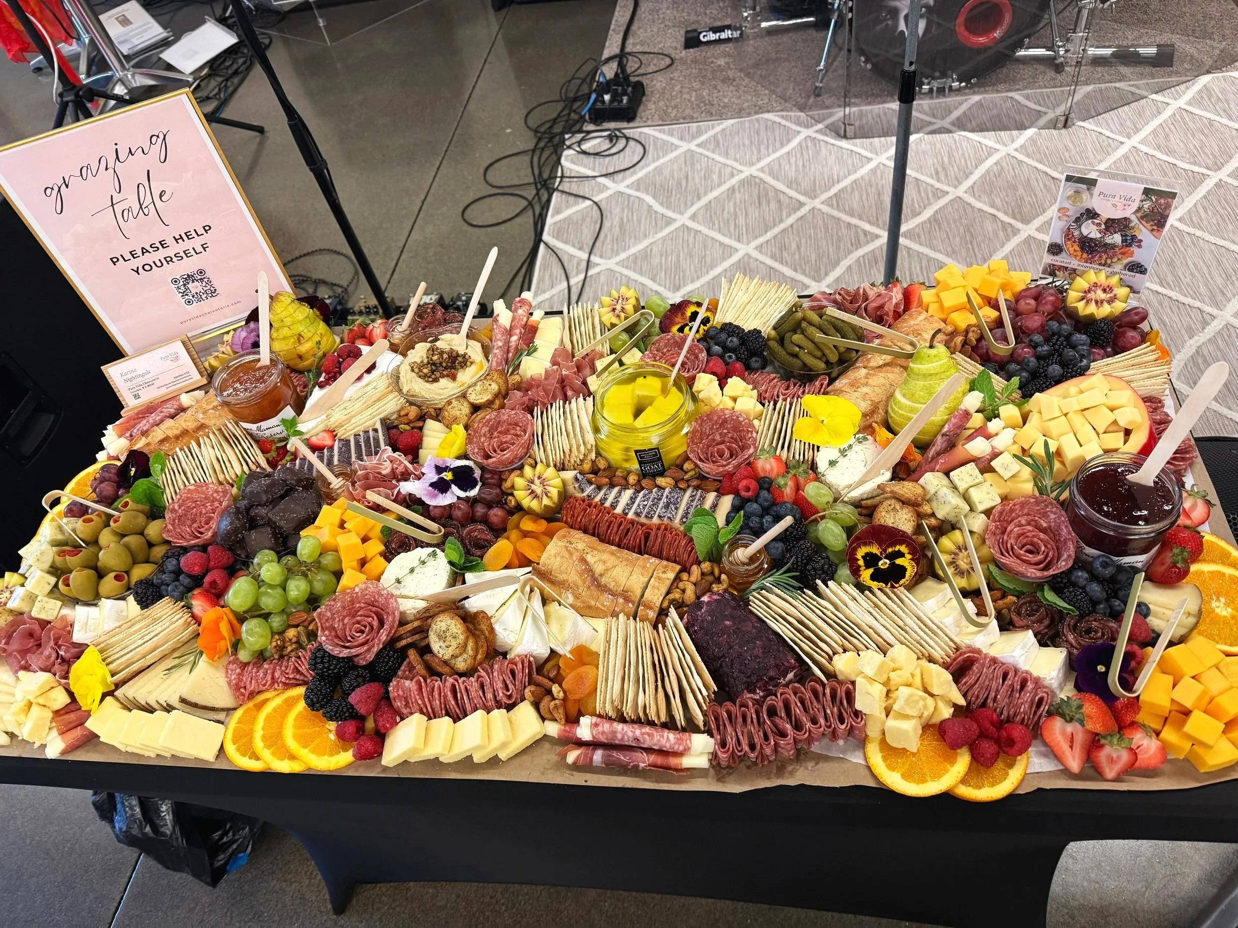 A large, lavish charcuterie board with cheese, meats, fruits, nuts, crackers, and condiments, decorated with edible flowers, on a black table at an event. Signage indicates it is an 'amazing table' for self-help, with one jar of honey or jam, a jar of olive oil, and small bowls of jams or spreads.