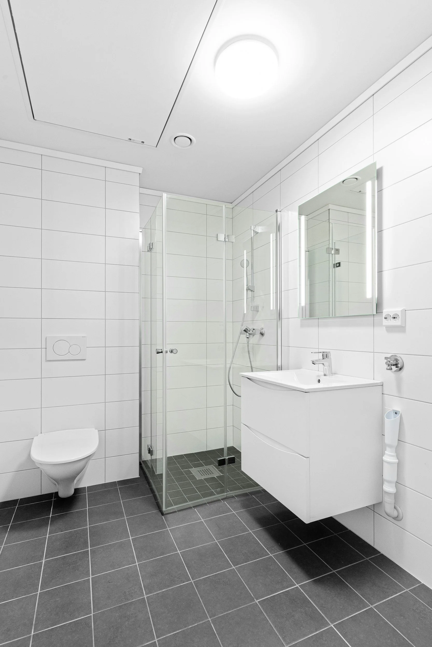 Valkoinen kylpyhuone, jossa wc, suihkukaappi ja valkoinen pesuallas lavuaarilla, peili ja valoisat valaisimet seinällä.