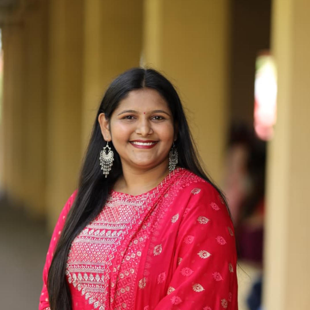 A woman smiling, wearing a pink ethnic dress with silver earrings, standing in a corridor with yellow walls.