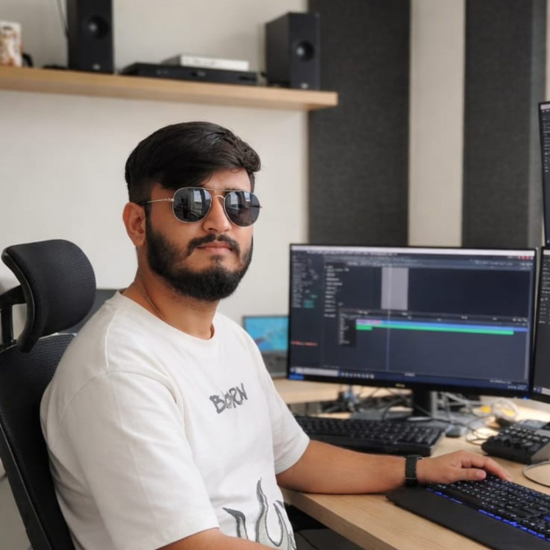 Man with dark hair and beard wearing sunglasses and a white T-shirt sitting at a desk with multiple monitors, working on video editing software.
