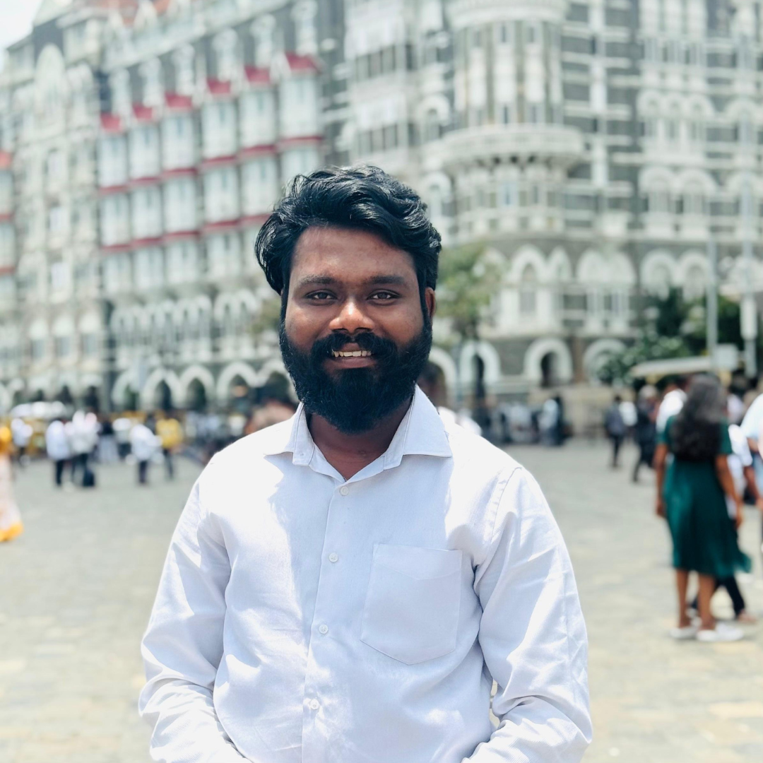 A man with a beard and dark hair smiling outdoors in front of a busy city square with a large, ornate multi-story building in the background. He is wearing a white shirt.