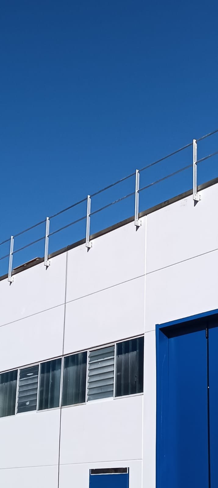 Facciata di un edificio moderno con pareti bianche, alcune finestre con lamelle di ventilazione e una grande porta blu, sotto un cielo blu senza nuvole.