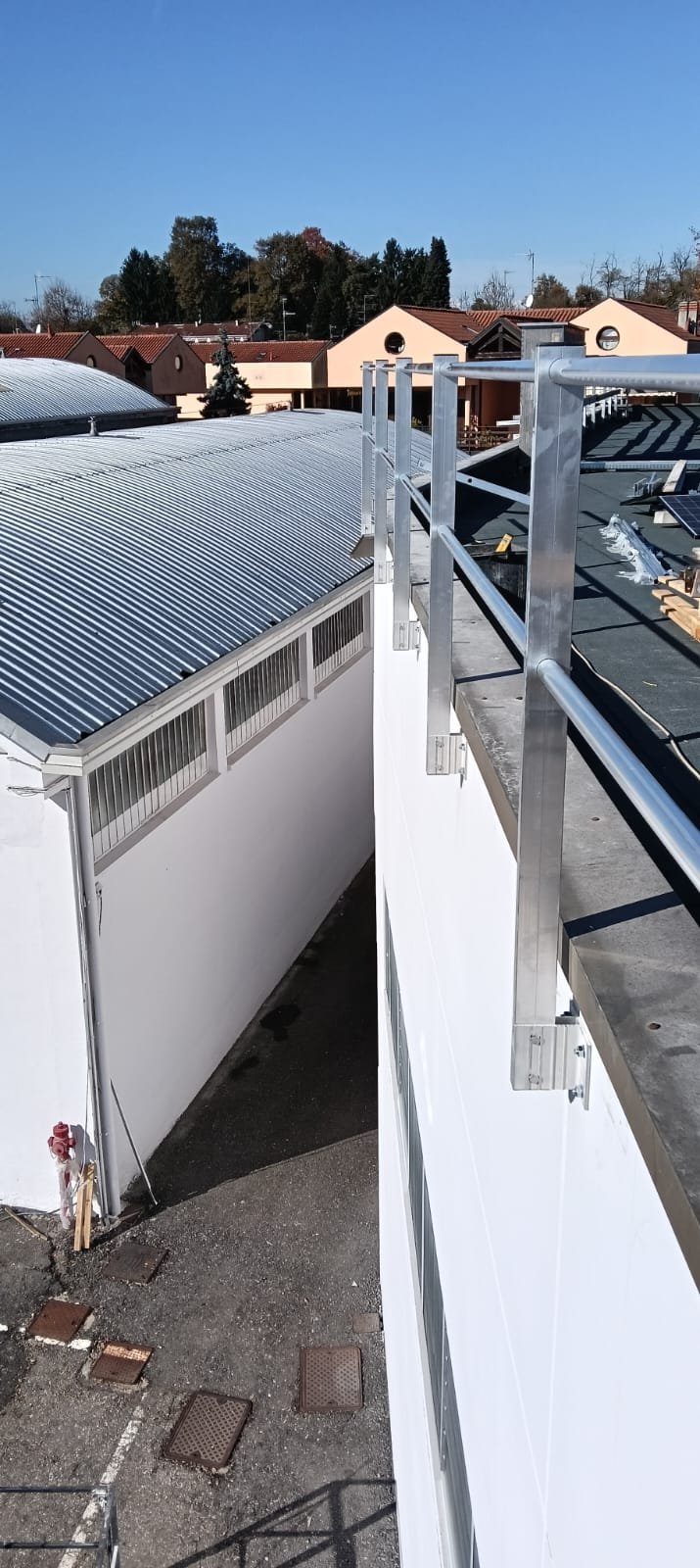 Vista di un tetto industriale con pannelli solari e un parapetto di sicurezza in alluminio, con alcune case e alberi sullo sfondo.