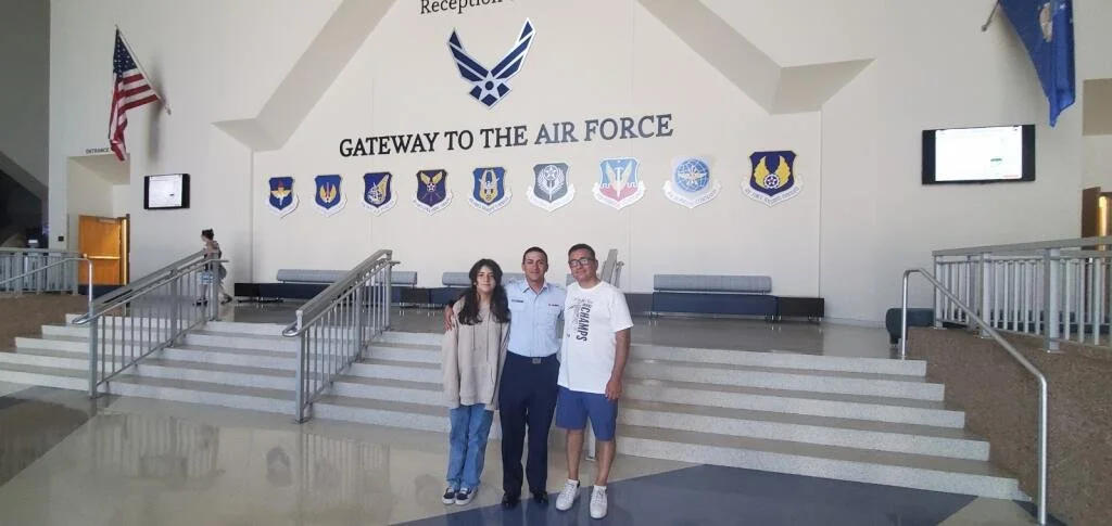 Three people standing in front of a mural that says 'Gateway to the Air Force'. The mural features various military insignias. An American flag is visible on the left side of the image.