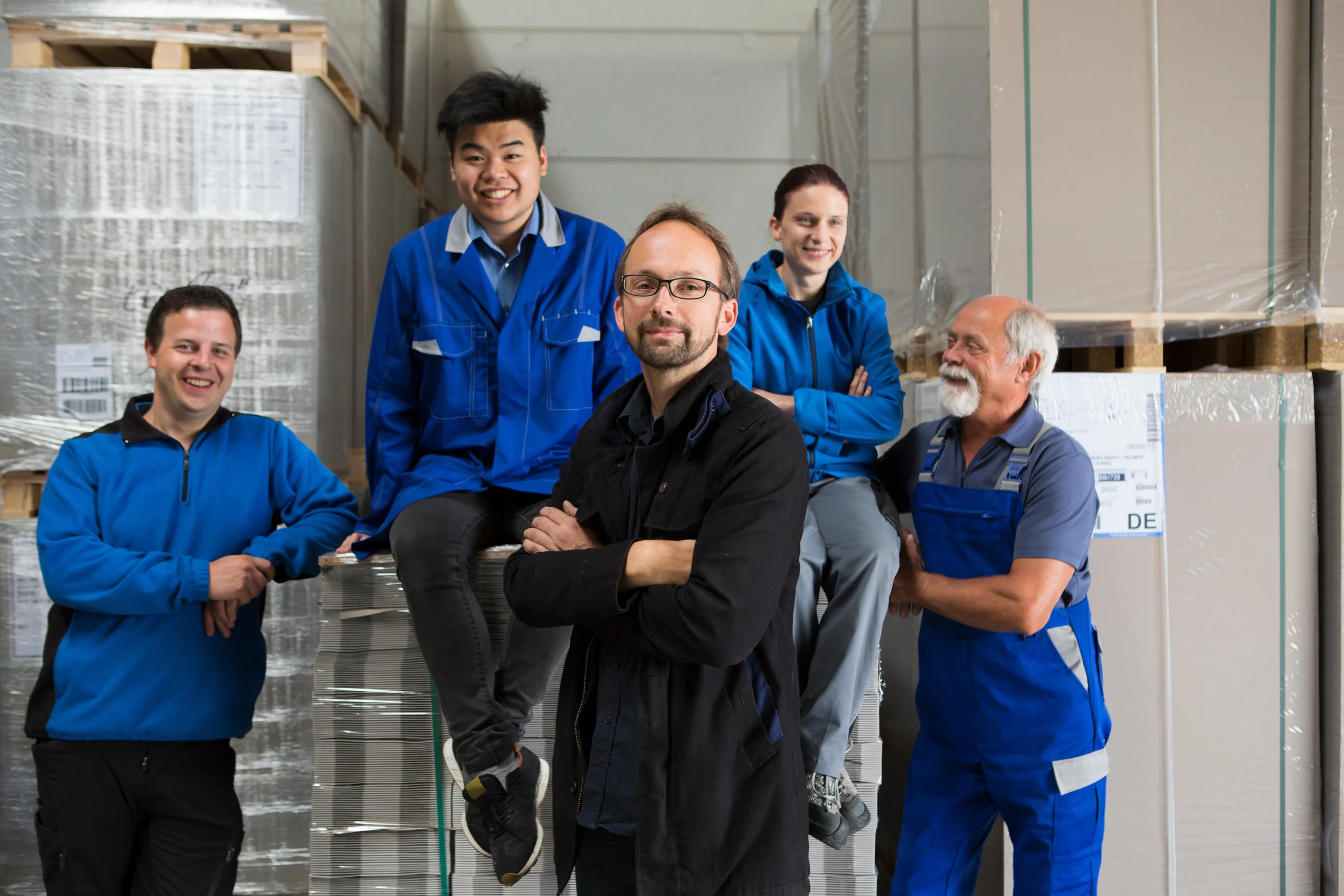 Grupp von sechs Menschen in blauen Arbeitskleidung in einem Lagerraum mit Paletten und Kartons.