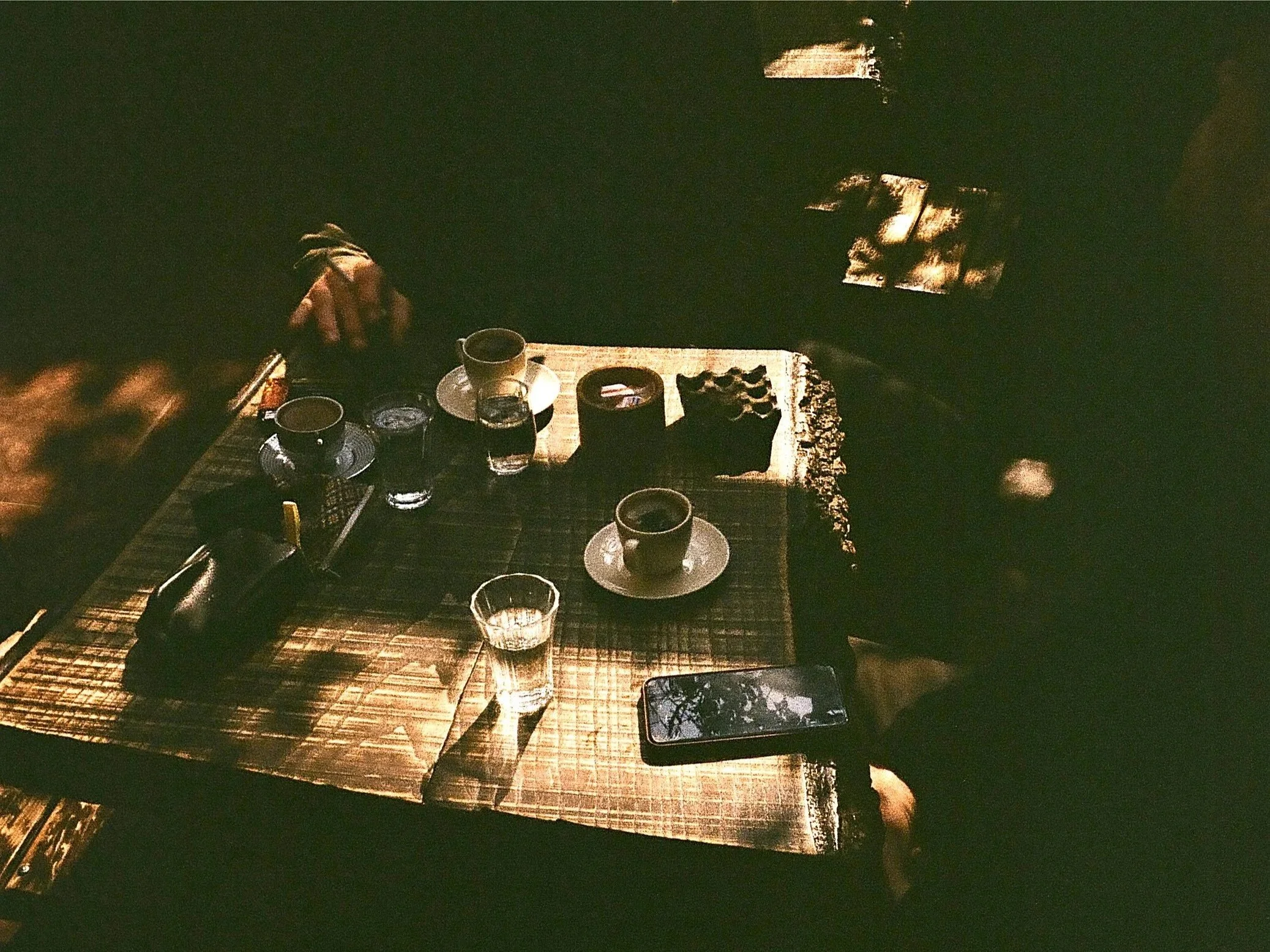 A wooden table with two cups of coffee, three glasses of water, a smartphone, and some small bowls. Hands are visible on either side of the table in a dimly lit setting.