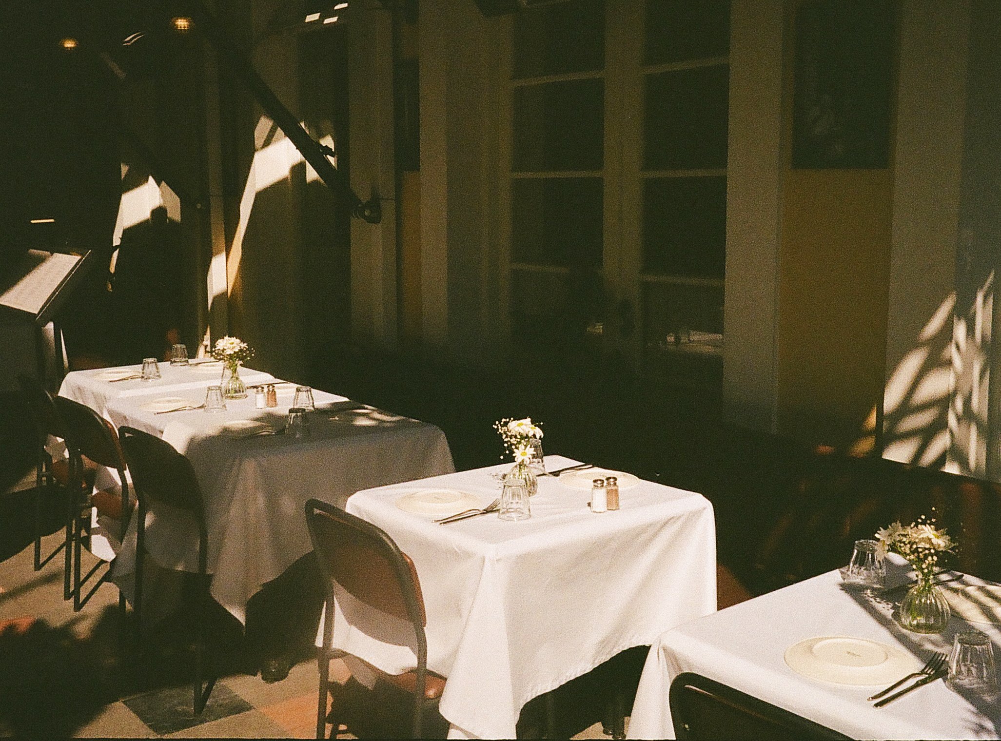 An empty restaurant with three tables covered with white tablecloths, small flower arrangements, glasses, and cutlery. Shadows from window frames create patterns on the wall and tables, with warm lighting.