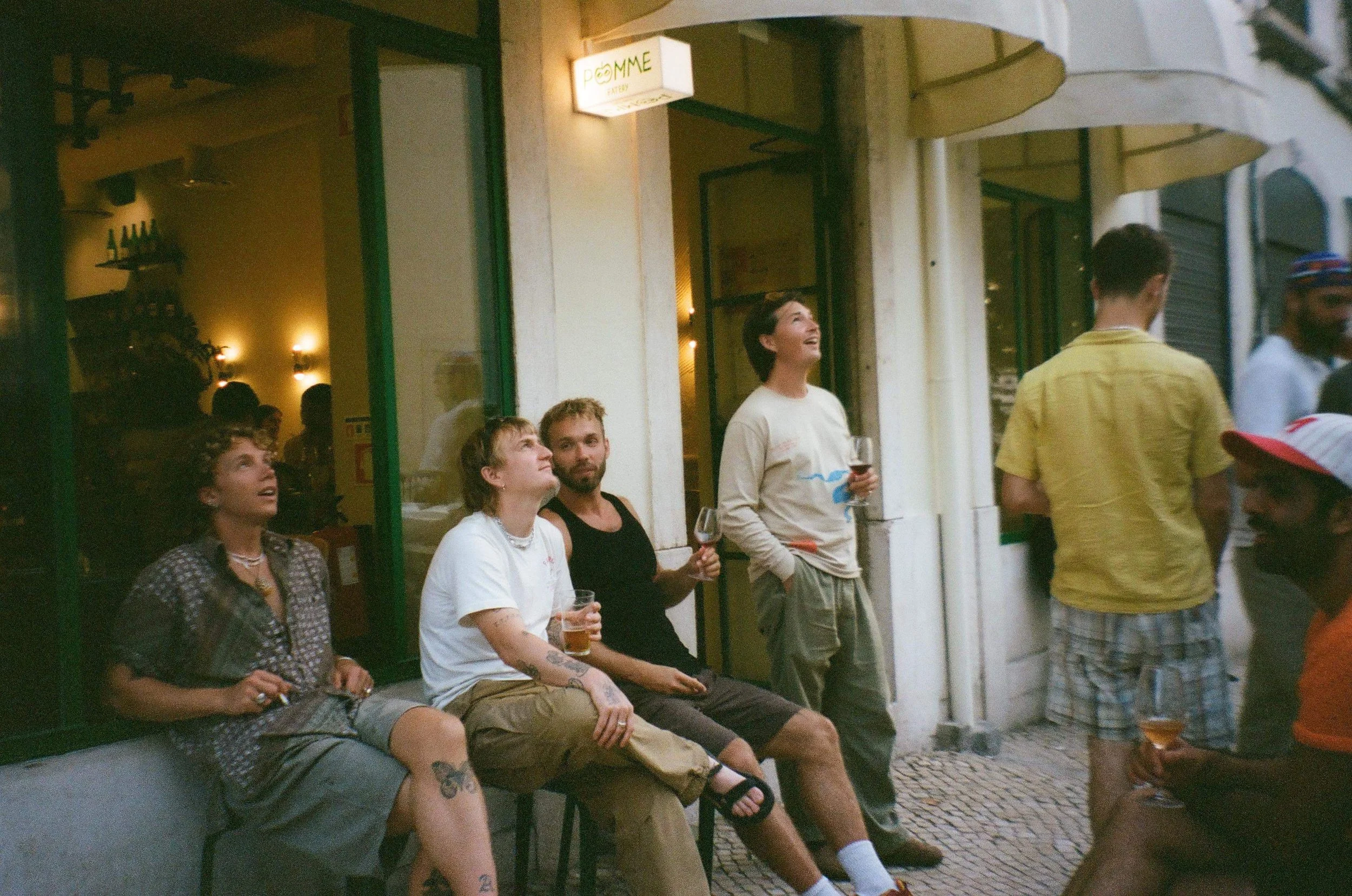 Group of people sitting and standing outside a bar or café, some holding drinks, engaging in conversation and enjoying the evening.
