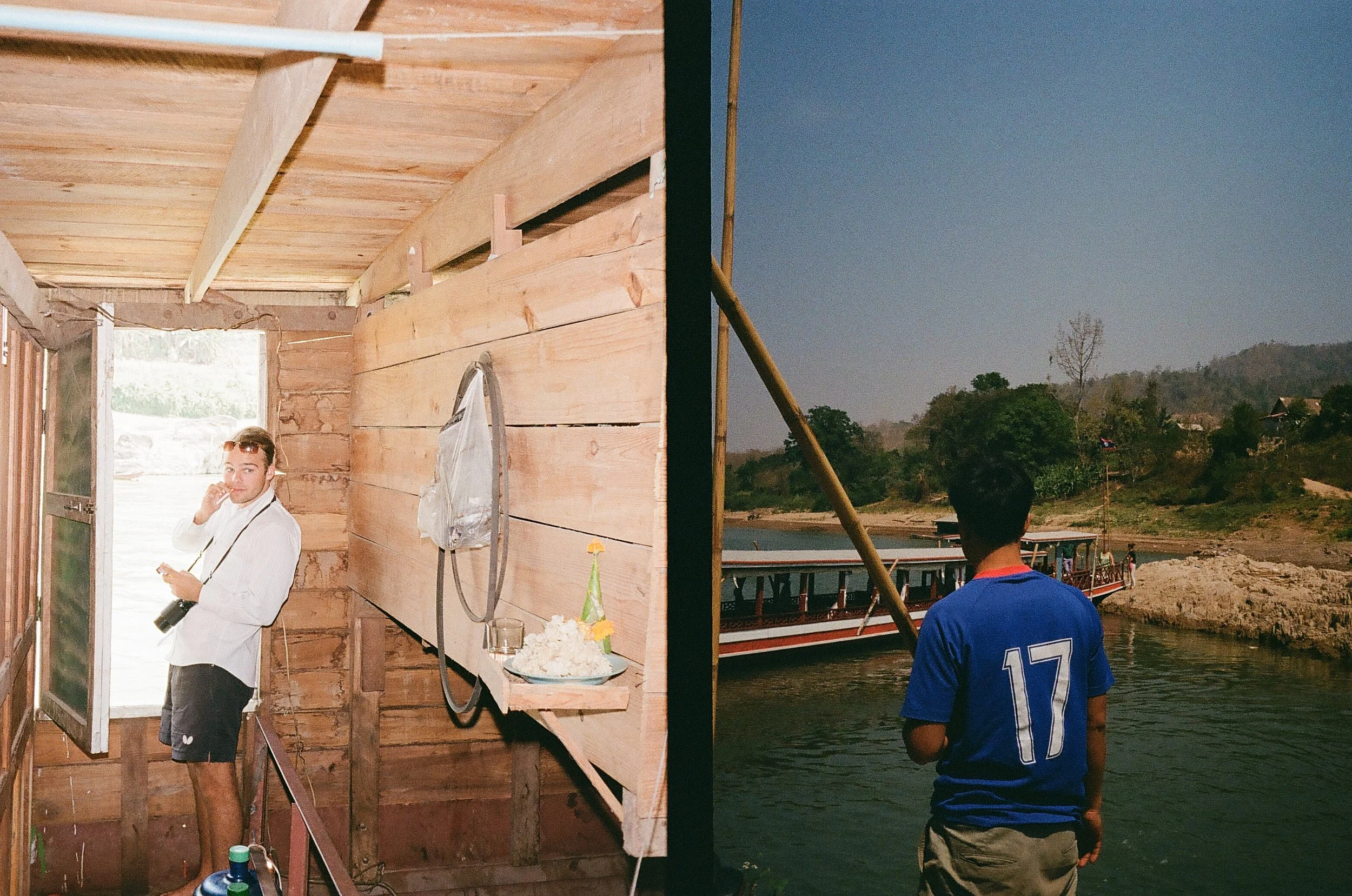 A person standing inside a rustic wooden structure with an open door, holding a camera. Another person is standing outside near a river, looking at a boat.