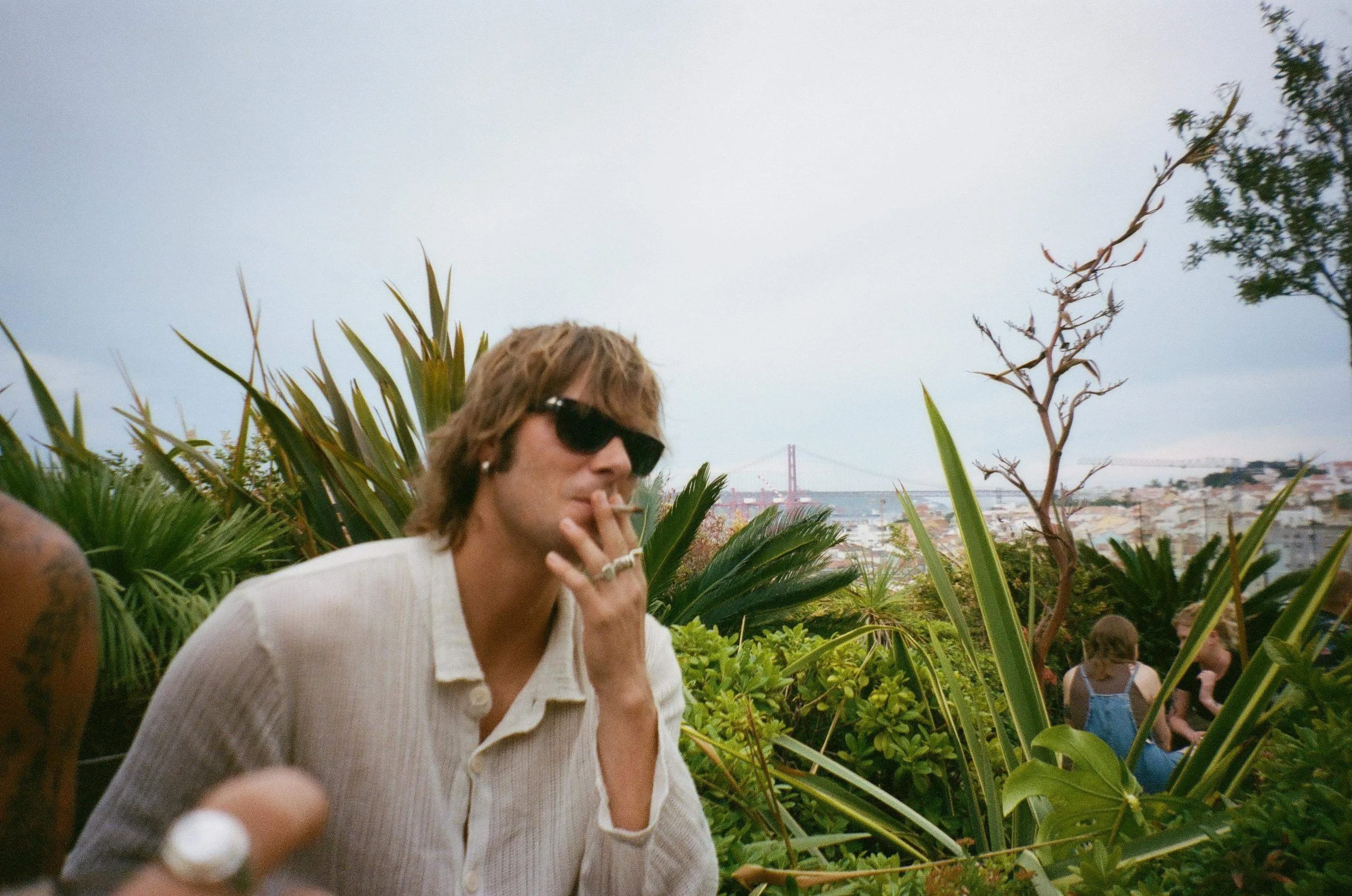 A person with light brown hair, wearing dark sunglasses and a beige shirt, smoking outdoors among lush green plants. In the background, the Golden Gate Bridge and a cityscape can be seen under a cloudy sky.