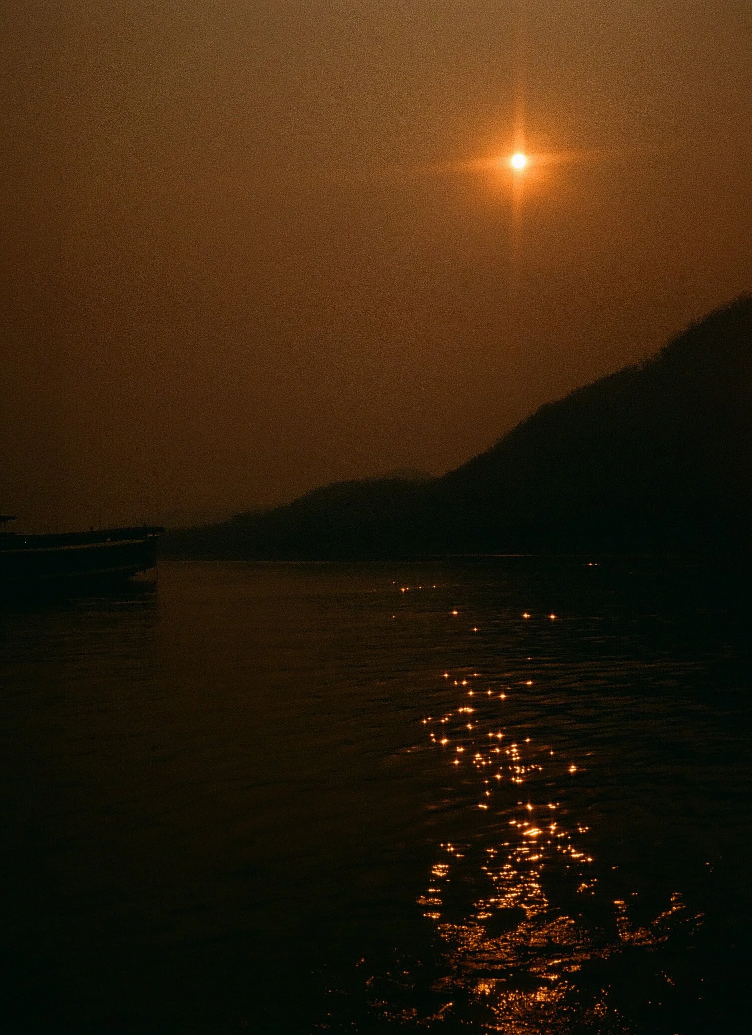 A nighttime scene of a river with a boat on the left side, a mountain on the right, and a bright moon shining in the sky. The moon's reflection creates a path of light on the water.