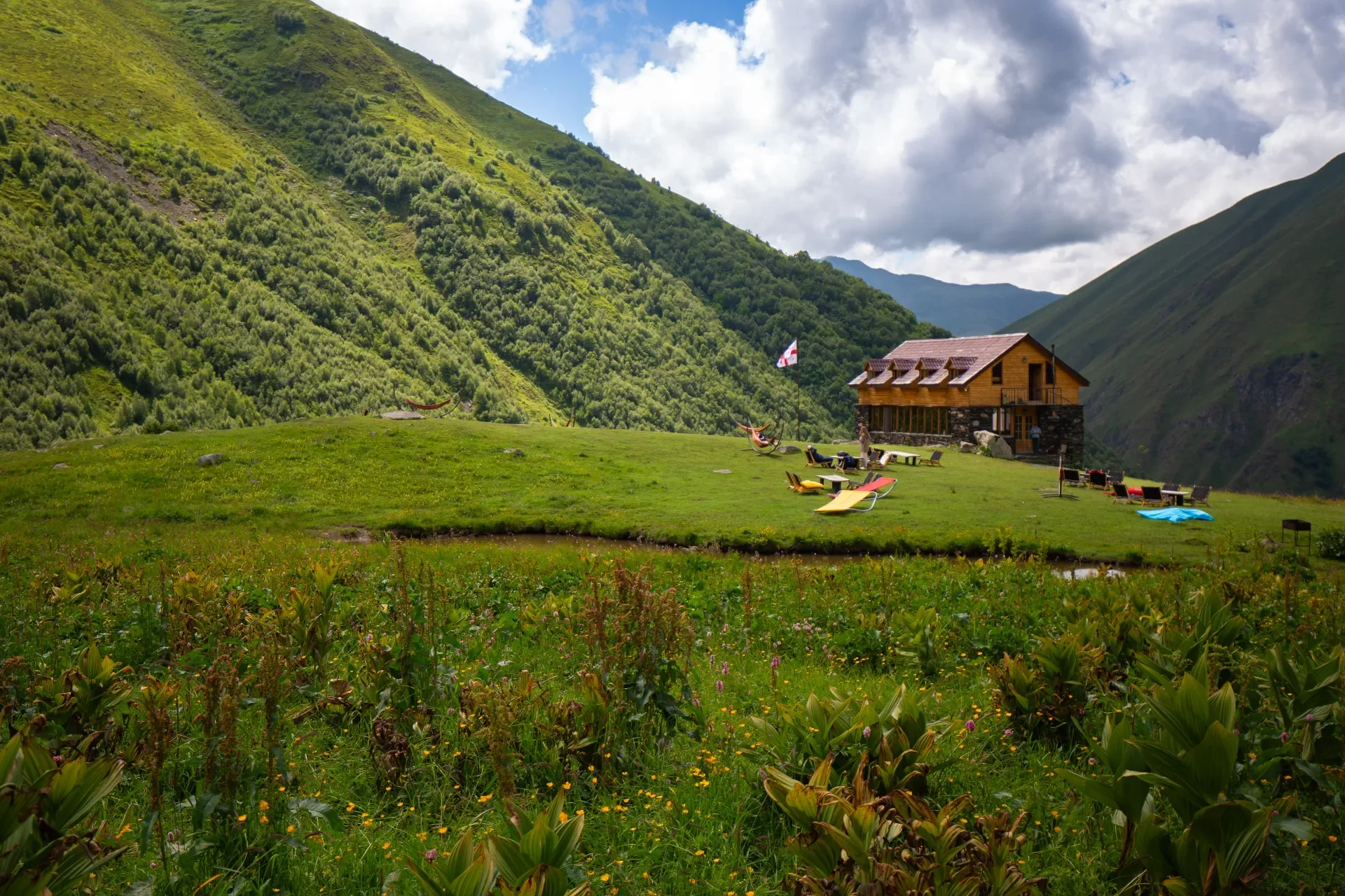 A scenic mountain landscape with a wooden house, green hills, and colorful outdoor furniture and hammocks on the grass.