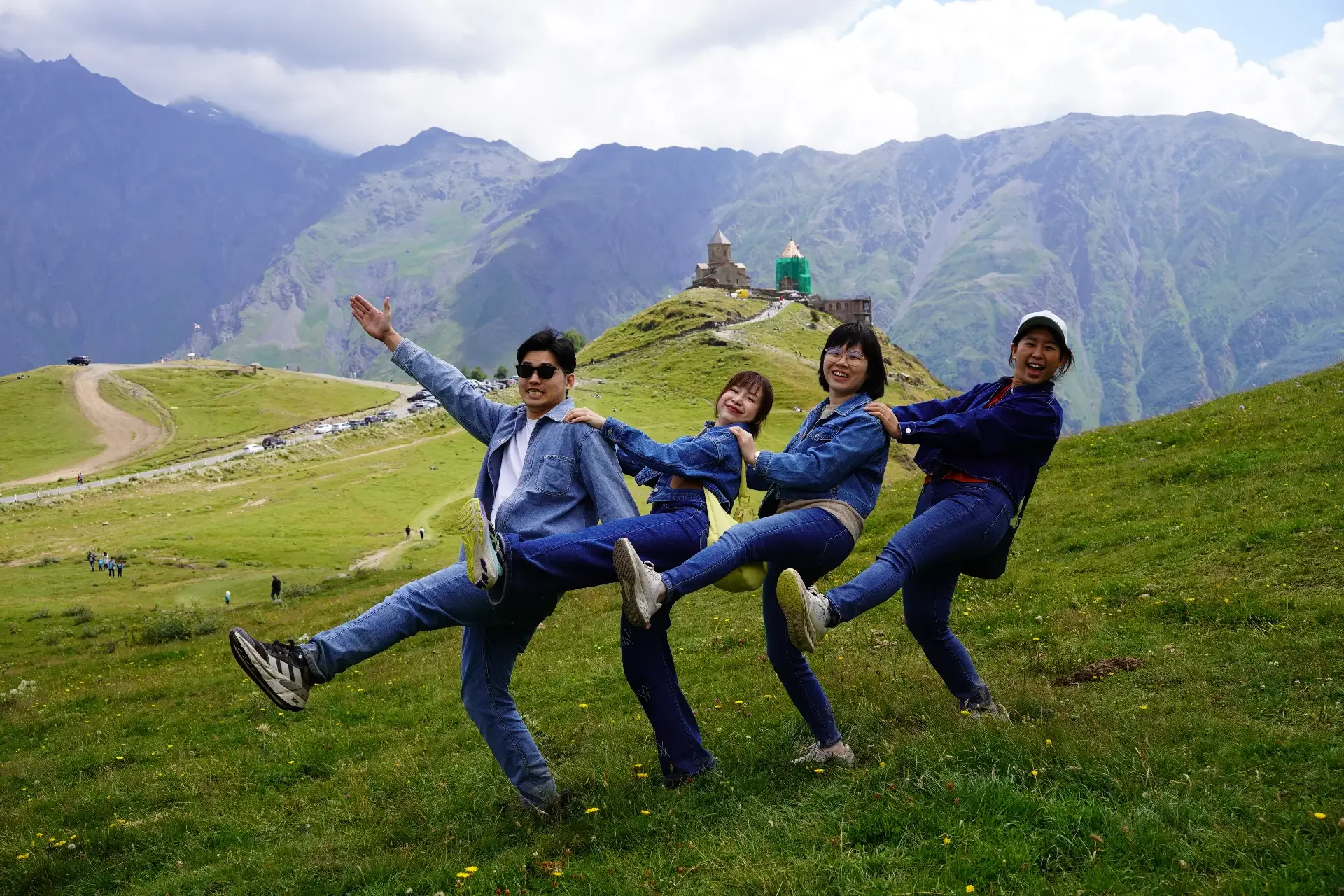 Four friends in denim outfits playfully pose in a line on a green mountain hillside with a small church and mountain range in the background.