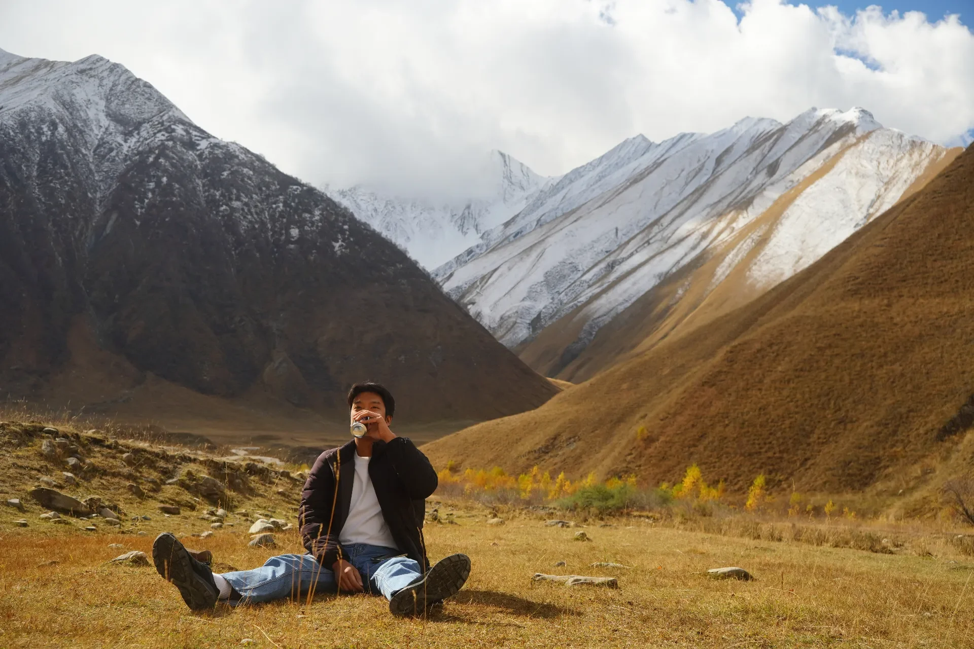 A person sitting on the grass in a valley, drinking a can, with snow-capped mountains in the background.