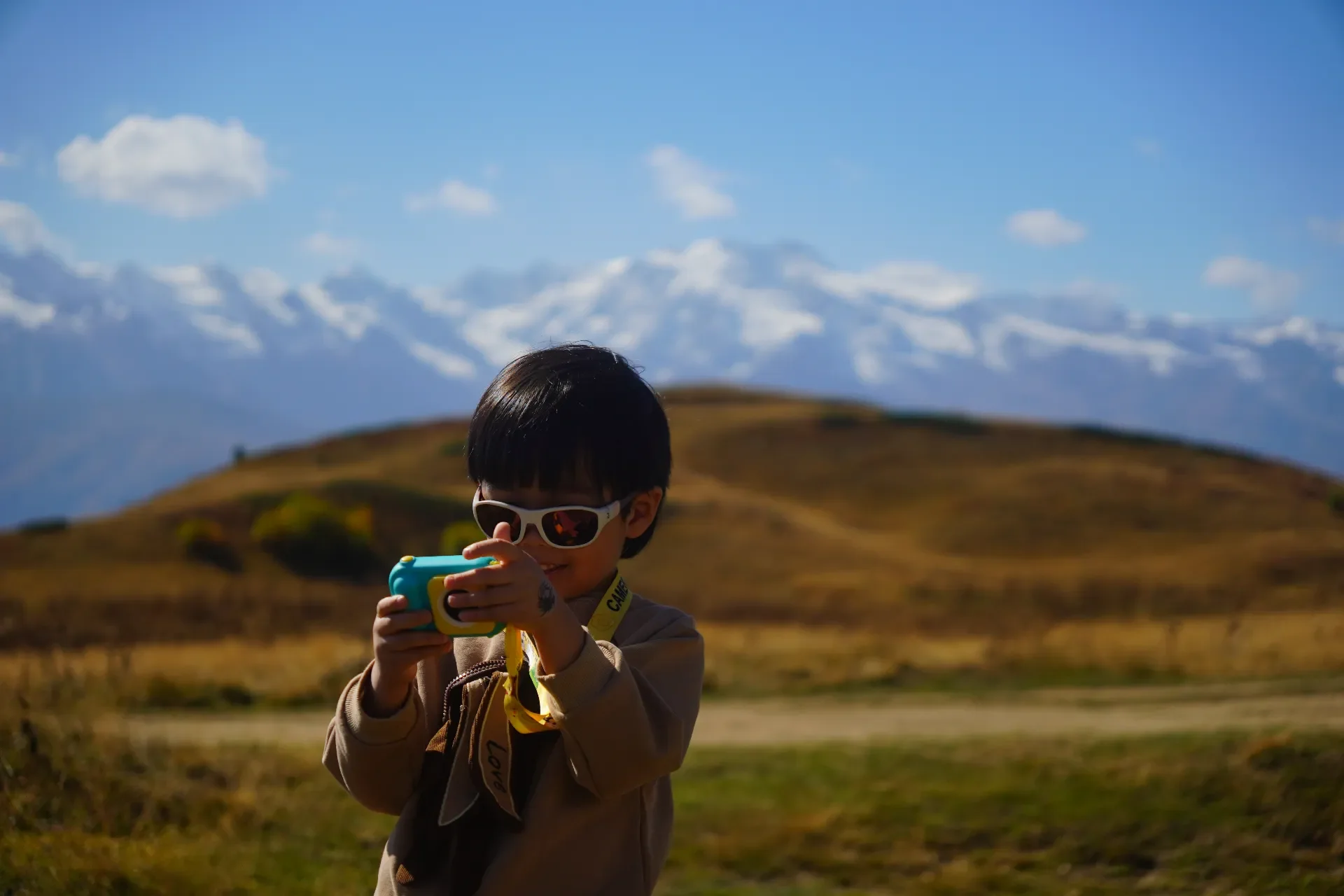 A smiling young boy wearing sunglasses and a beige jacket, holding a colorful toy camera, standing in a scenic outdoor landscape with rolling hills and snow-capped mountains in the background.