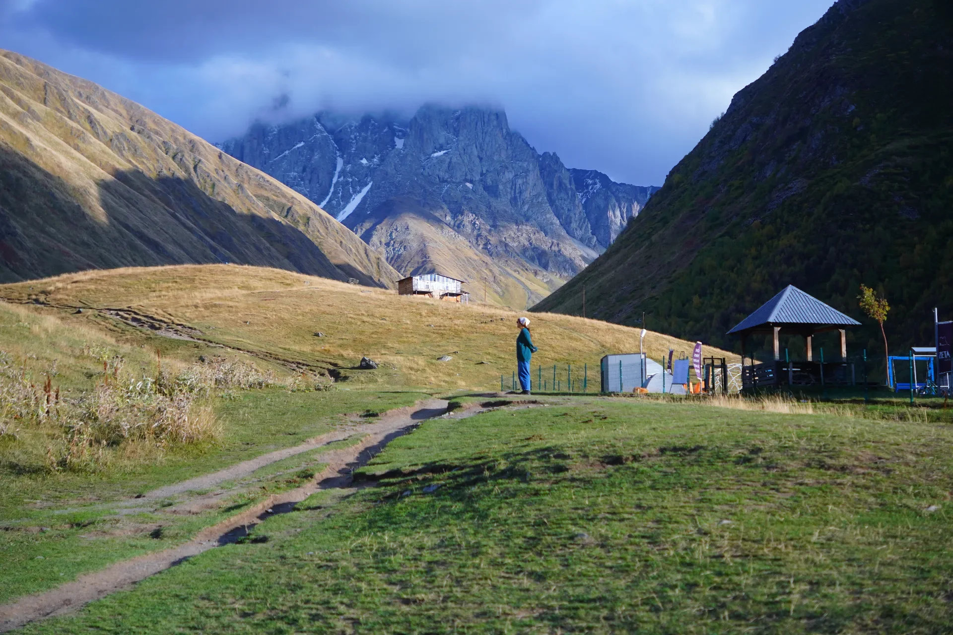 A person standing on a grassy path in a mountainous landscape with steep green slopes and snow-capped peaks in the background. There are small buildings and a pavilion nearby.