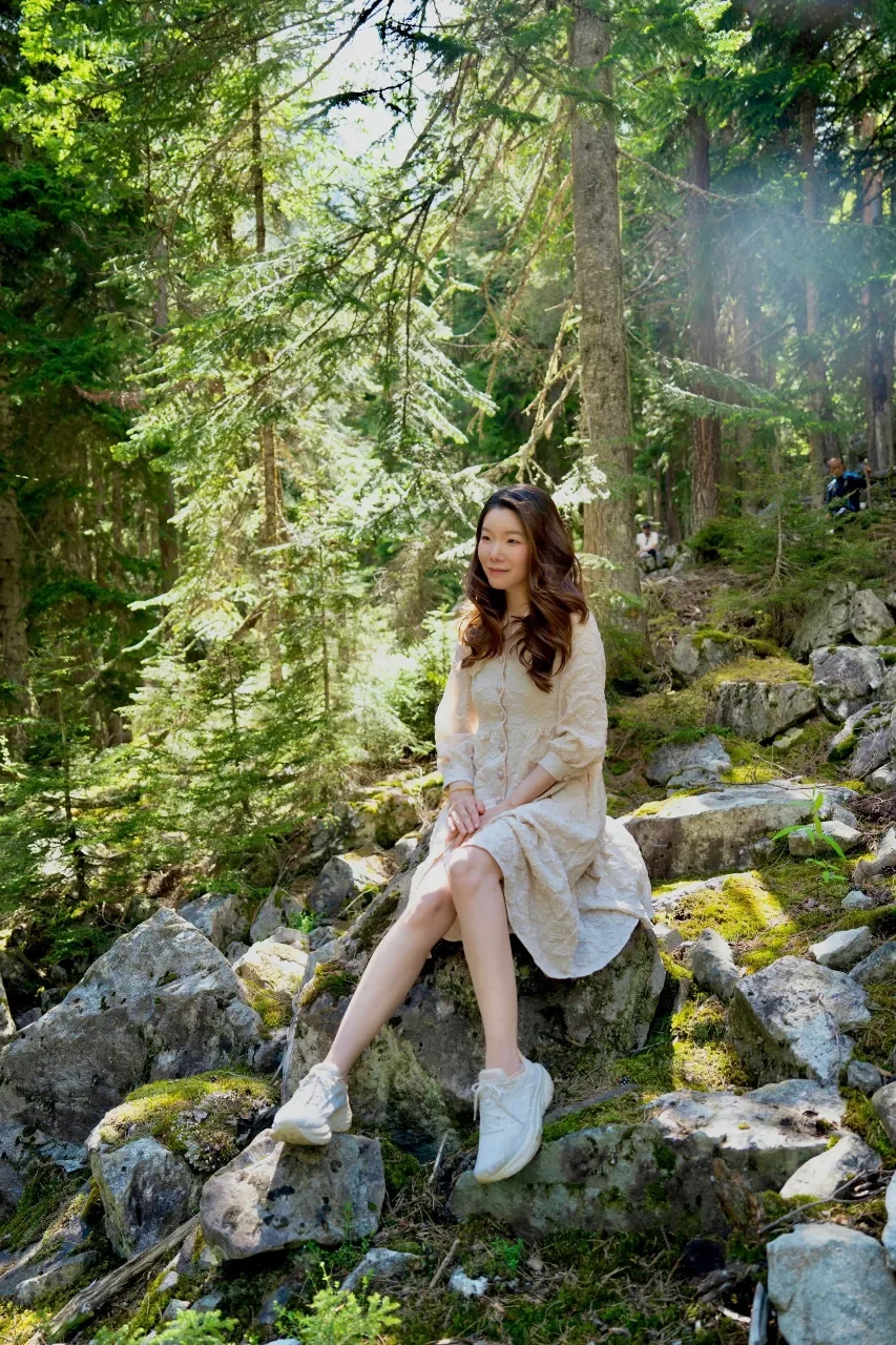 A young woman with curly brown hair sitting on a moss-covered rock in a lush, green forest surrounded by tall trees and rocks, wearing a cream-colored dress and white sneakers, smiling softly.