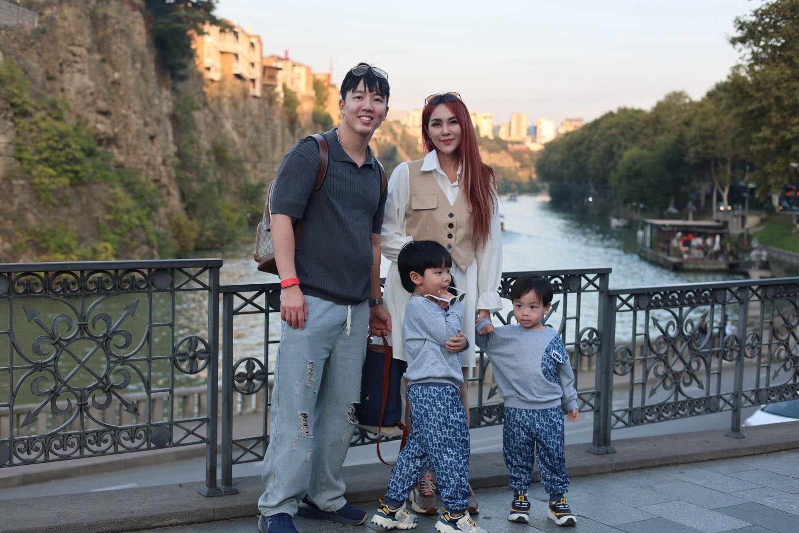 A family of four standing outdoors near a river with trees and buildings in the background, posing for a photo. The father and mother stand behind two young children, all smiling.