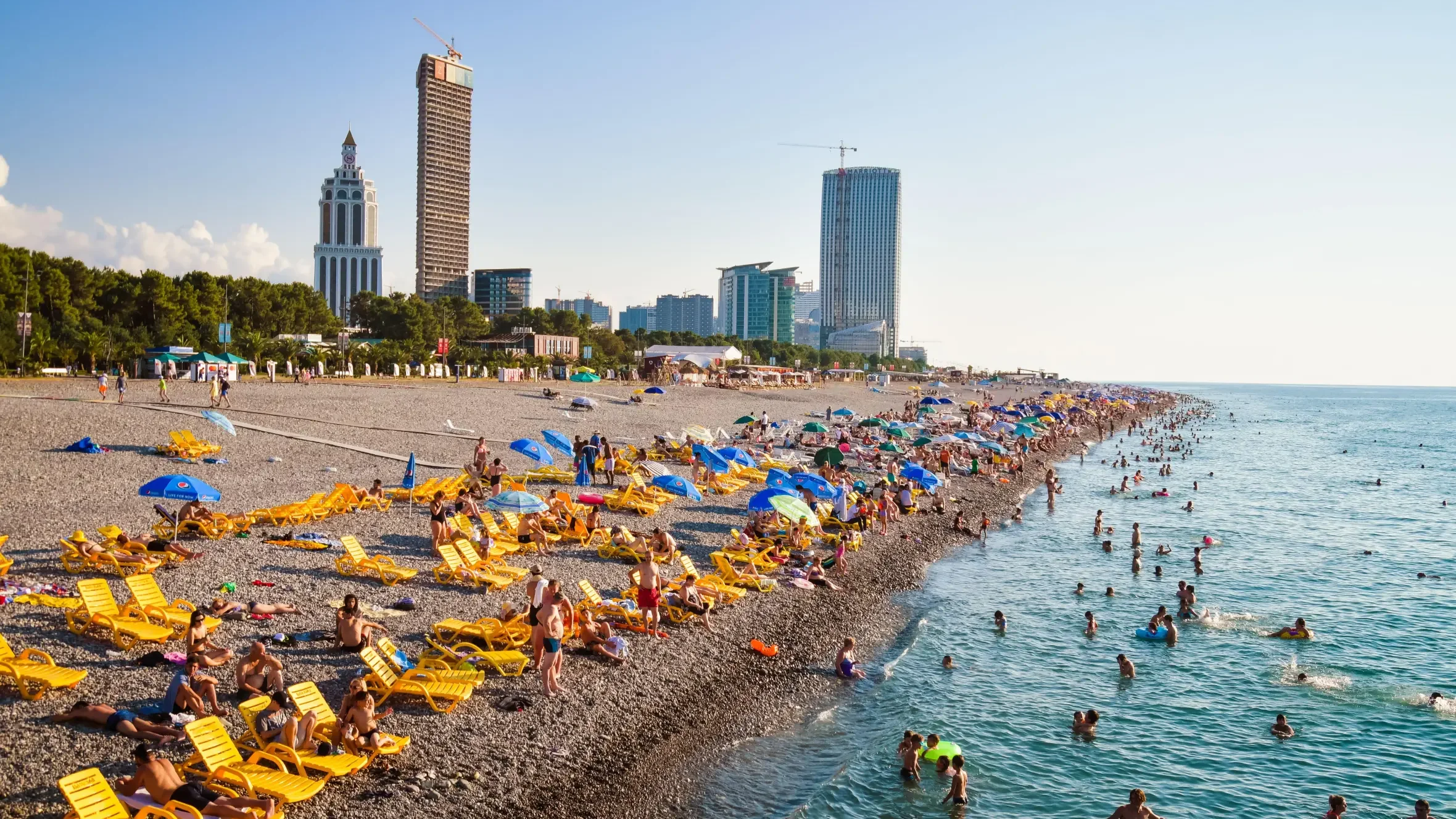 Crowded beach with many yellow lounge chairs and umbrellas, with people swimming and relaxing by the ocean, and tall buildings in the city skyline in the background.