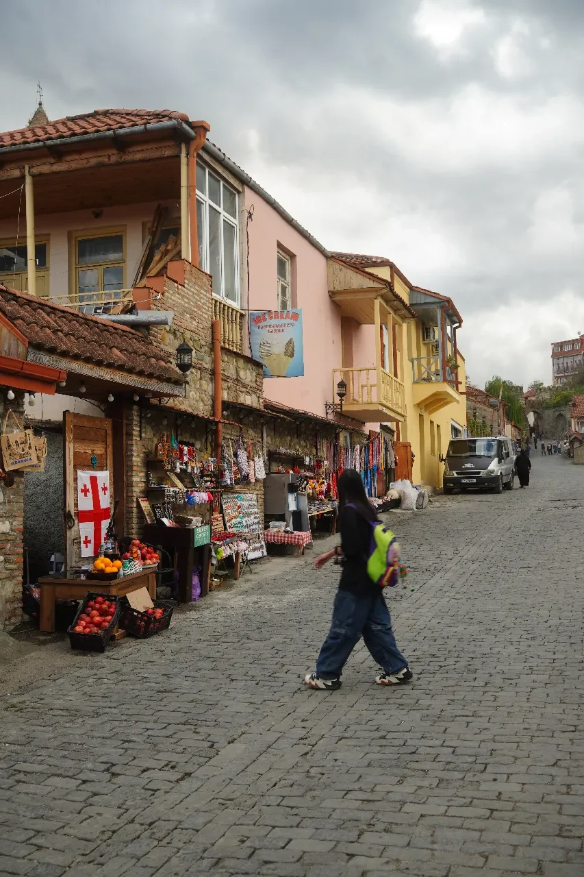 A cobblestone street in a hillside town with colorful buildings, a girl in dark clothing and a green backpack walking past market stalls with fruit and souvenirs, cloudy sky overhead.