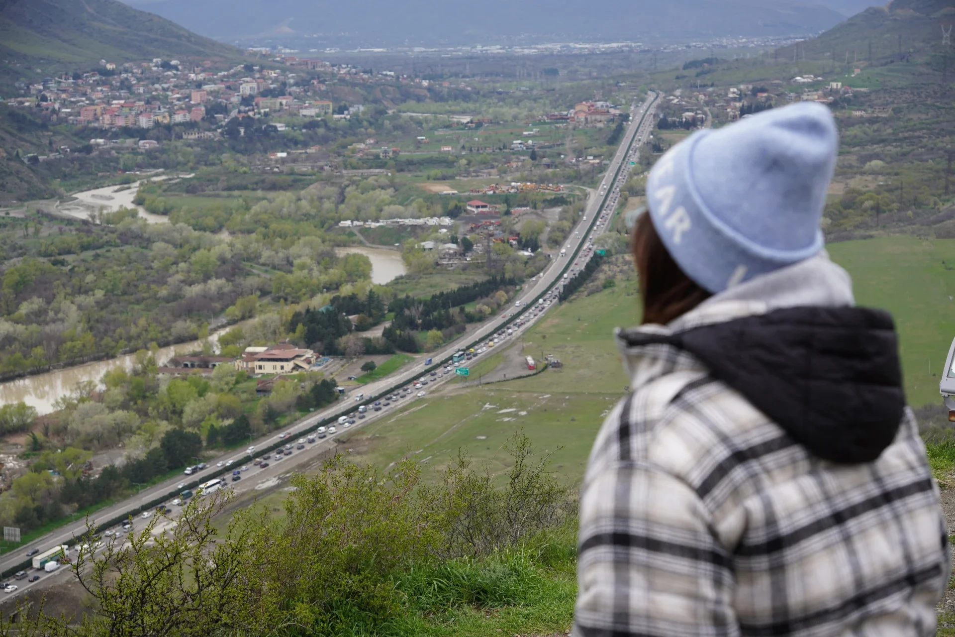 A person wearing a light blue hat and a plaid jacket sitting on a grassy hill, overlooking a valley with a river, a highway with traffic, and a town with many buildings in the distance.