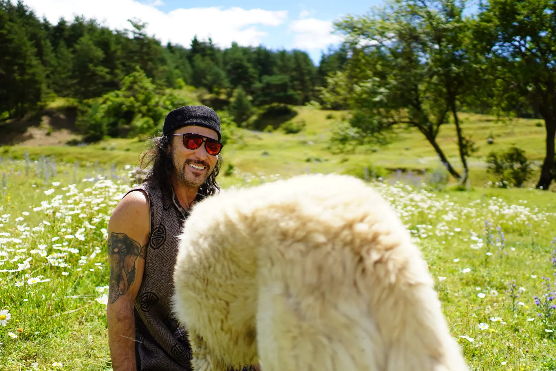 A man with tattoos, sunglasses, and a black beanie smiling in a field of flowers with a large white dog in front of him, surrounded by green trees and hills under a partly cloudy sky.