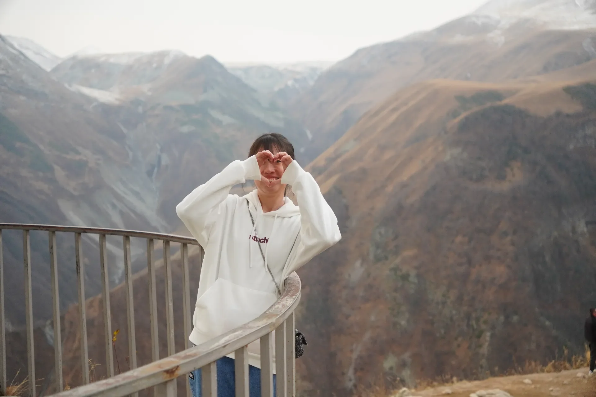 A person with a white hoodie standing on a mountain overlook, making a heart shape with their hands around their face, with mountain ranges in the background.