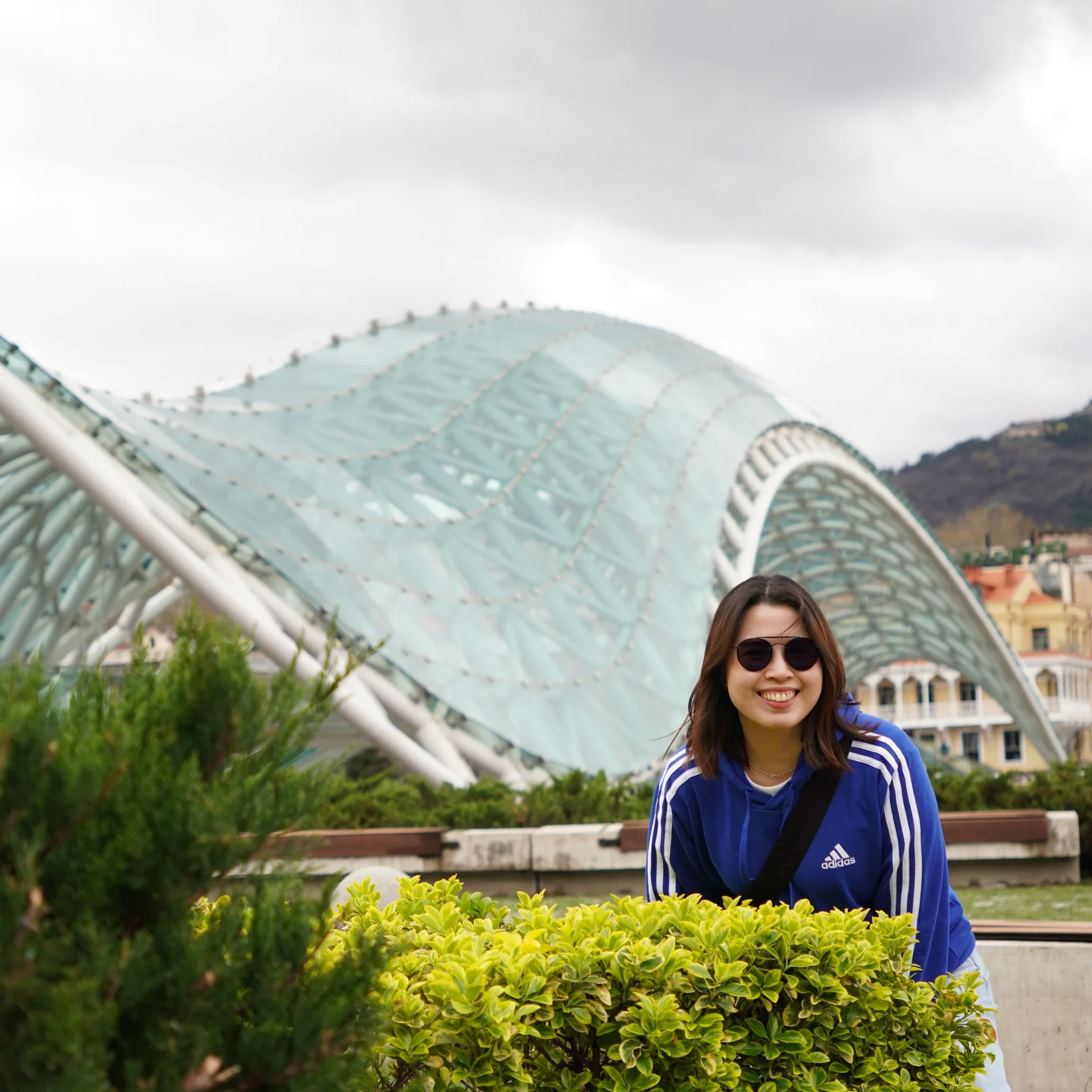Woman with brown hair and wearing sunglasses and a blue Adidas jacket smiling in front of green bushes and a modern architectural structure with a glass roof in the background.