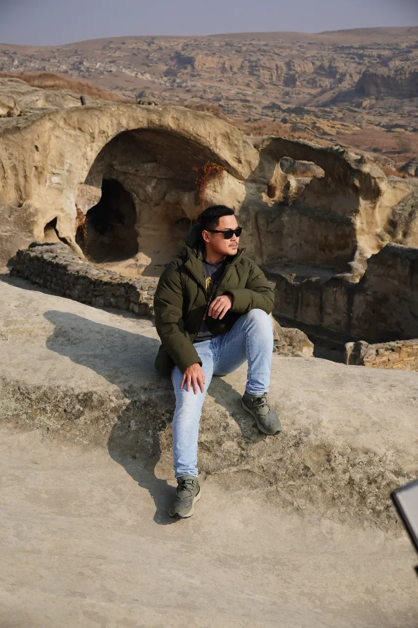 A man wearing sunglasses, a green jacket, and jeans sitting on a large rock in a rocky desert landscape with eroded rock formations and hills in the background.
