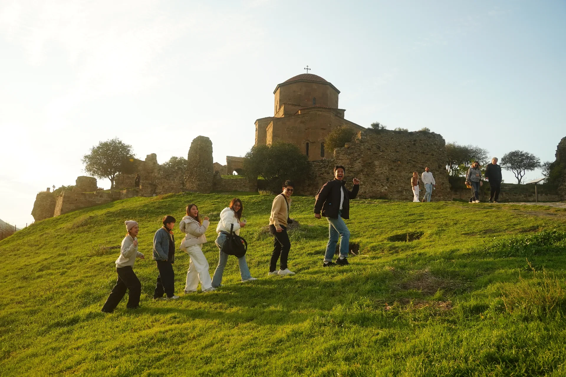 A group of people hiking up a grassy hill towards an ancient stone church and ruins, with a clear sky in the background.