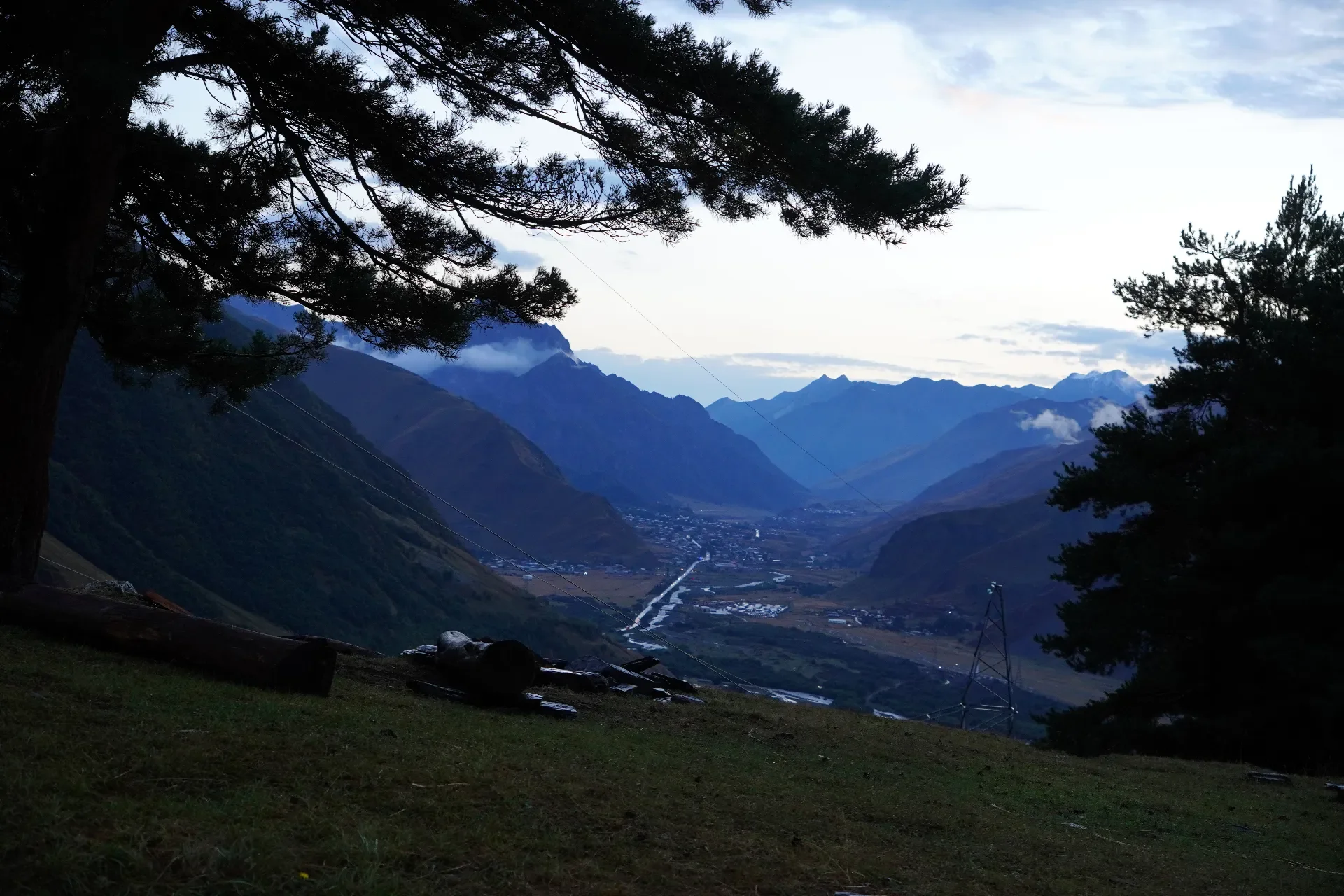 Mountain landscape at dusk with silhouette trees in the foreground and a valley with a small town and power lines in the distance.