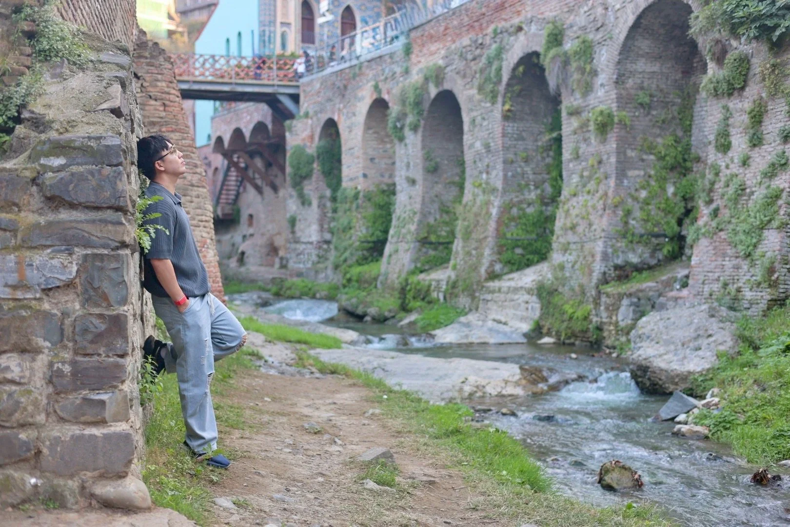 A young man leaning against a stone wall along a small river, with stone arches and buildings in the background, in a historic European town.