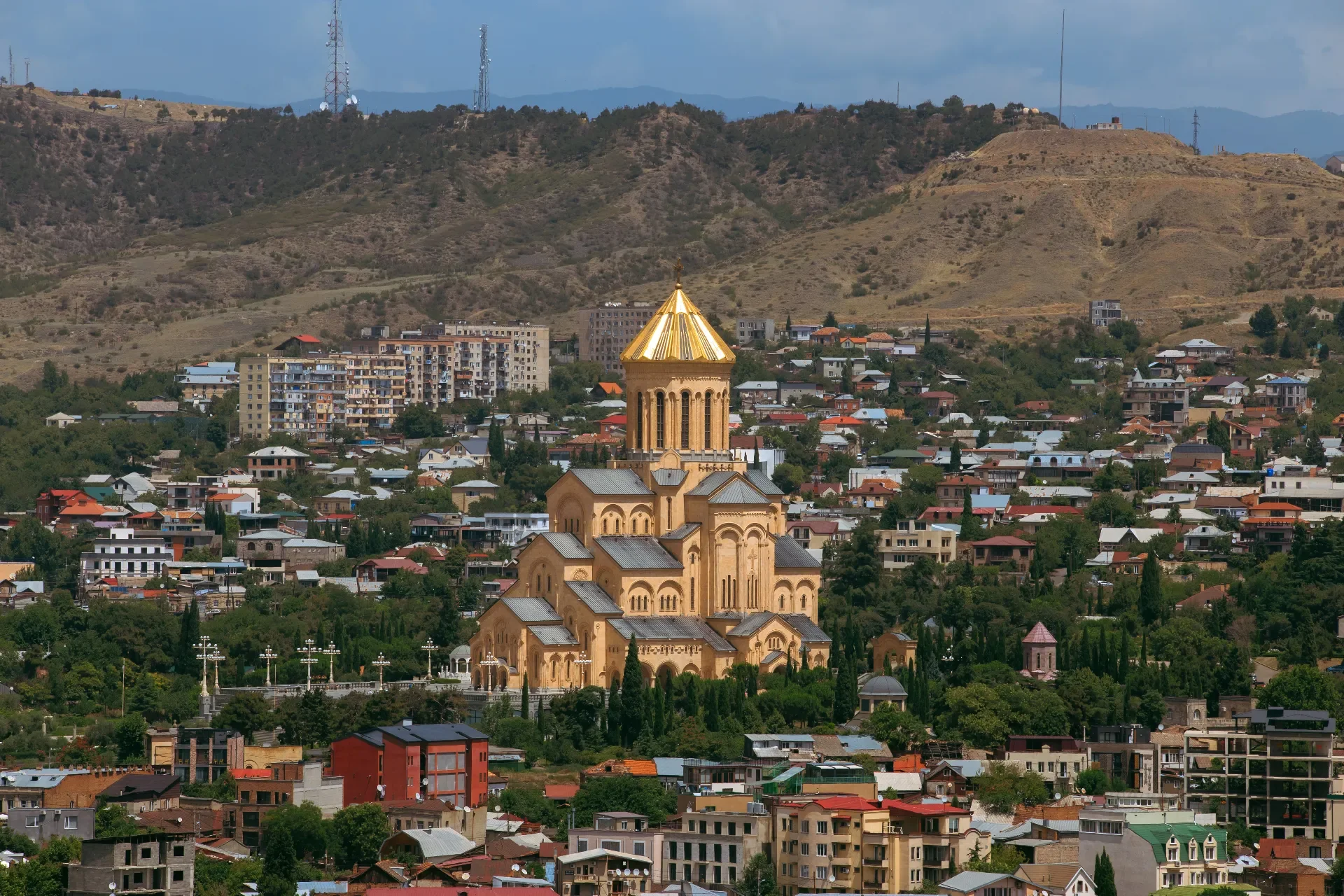 A cityscape with a large, golden-domed church in the center, surrounded by numerous residential buildings and green trees, with hills and mountains in the background.