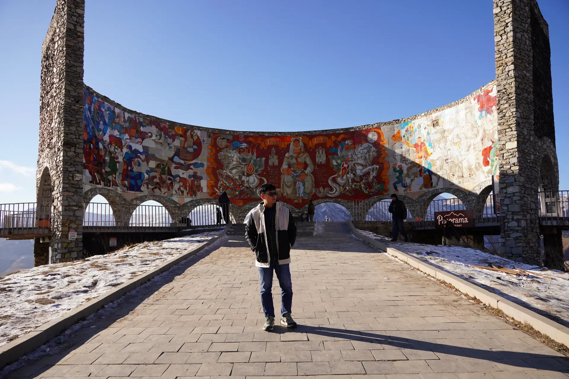 A person standing on a paved walkway in front of a stone structure with colorful murals, with mountains in the background and a clear blue sky.