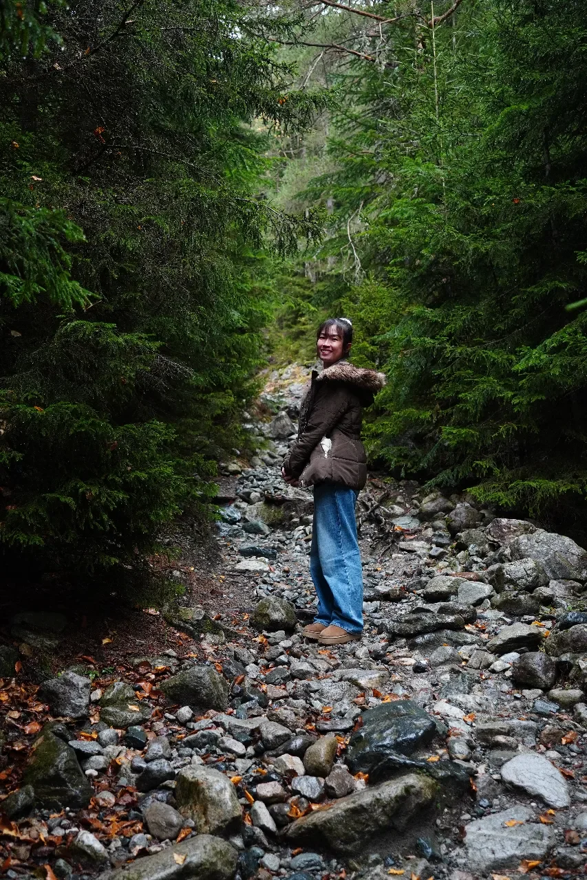 A woman standing on a rocky trail surrounded by dense green trees, smiling and looking back at the camera.