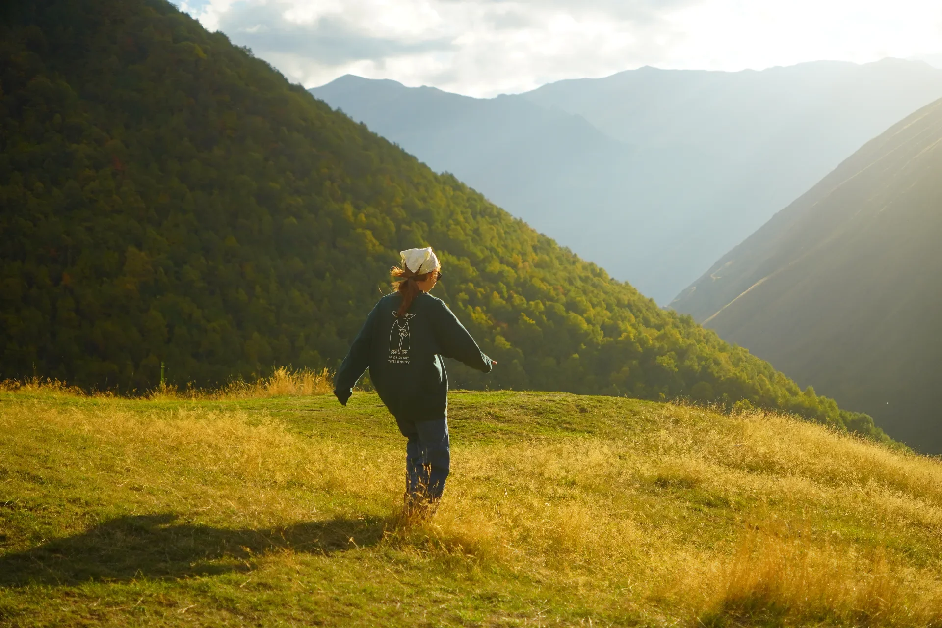 Person walking on a grassy hillside with mountains in the background under a partly cloudy sky.