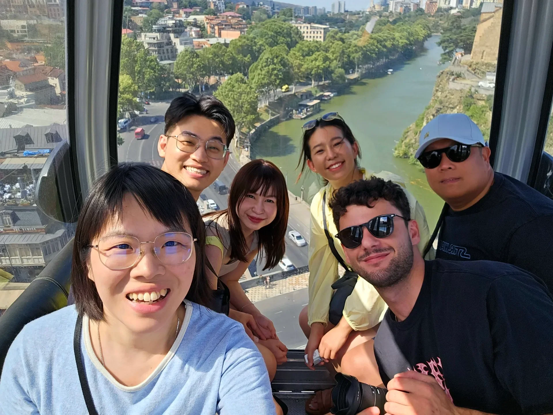 Group of six friends taking a selfie inside a Ferris wheel cabin with a city and river view behind them.