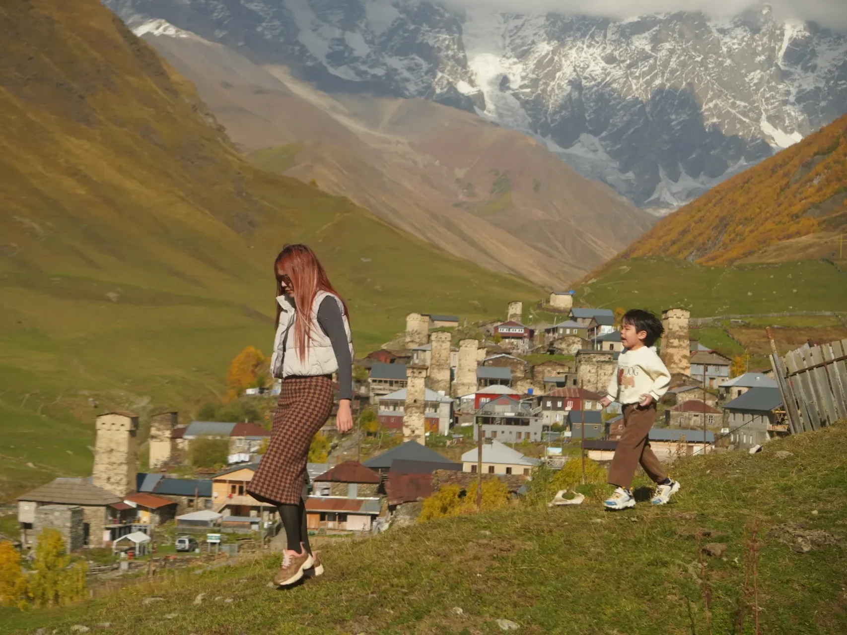 Two women are posing and smiling in a green meadow with mountains and snow in the background. One woman is making a gesture with her arm extended, pointing across the landscape.