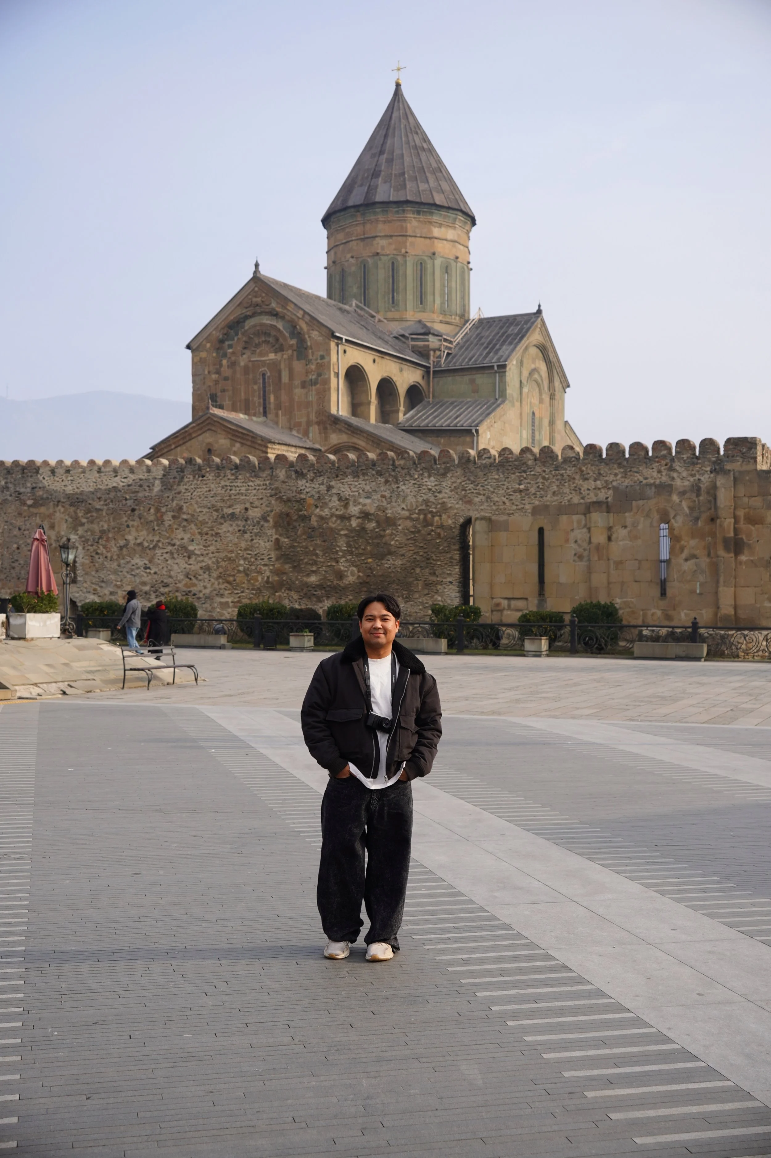 A man standing outdoors on a paved area with a historic stone church and castle in the background.