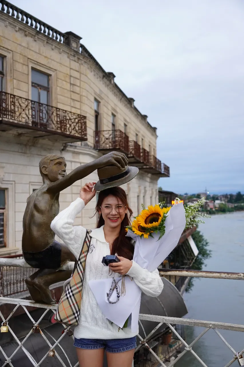 A young woman with glasses and long brown hair smiling while holding a bouquet of sunflowers and a camera, standing on a bridge near a river with historic buildings in the background.