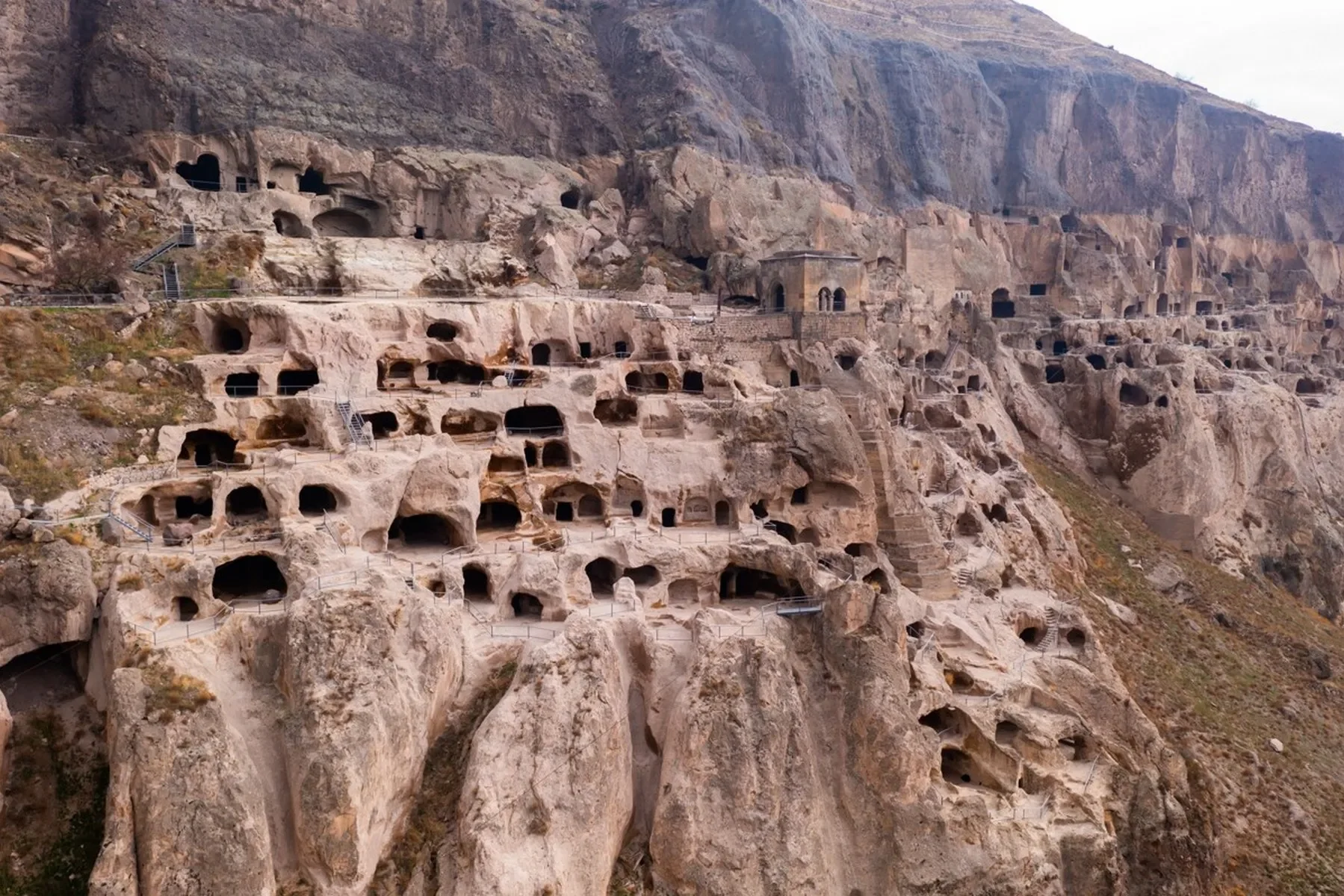 Ancient rock-cut tombs and dwellings carved into a mountainside with pathways and stairs connecting different levels.
