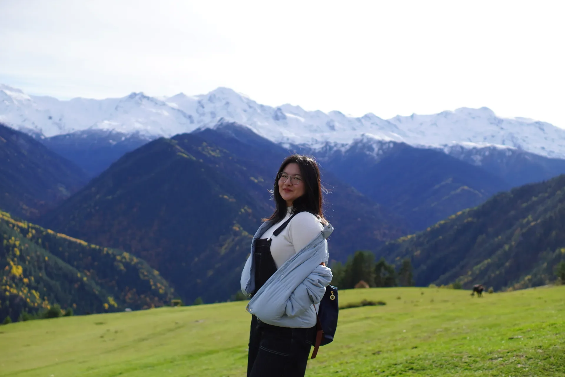 A woman with glasses and dark hair standing on a grassy field with mountains in the background. The mountains have some snow on their peaks, and the sky is bright.