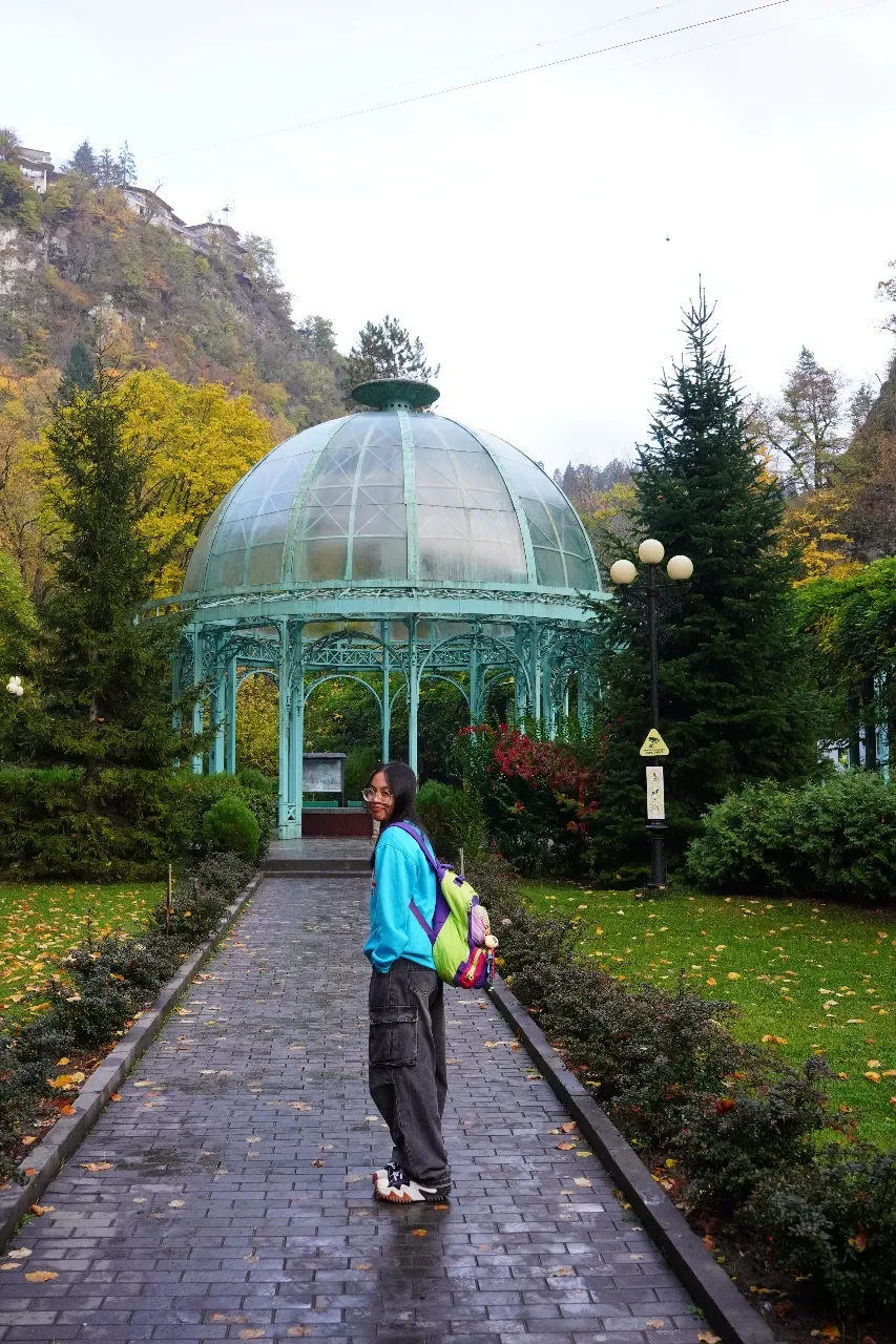 A young woman with glasses and a bright blue jacket standing on a wet brick pathway in a park with autumn leaves, green trees, and a glass-domed pavilion in the background.