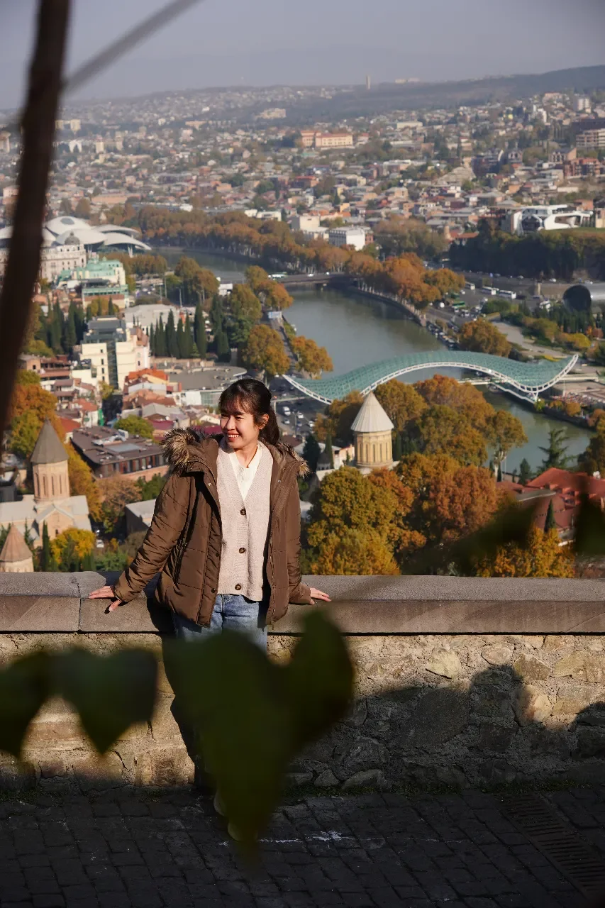 Young woman smiling and leaning on a stone railing with a cityscape, river, and bridge in the background.